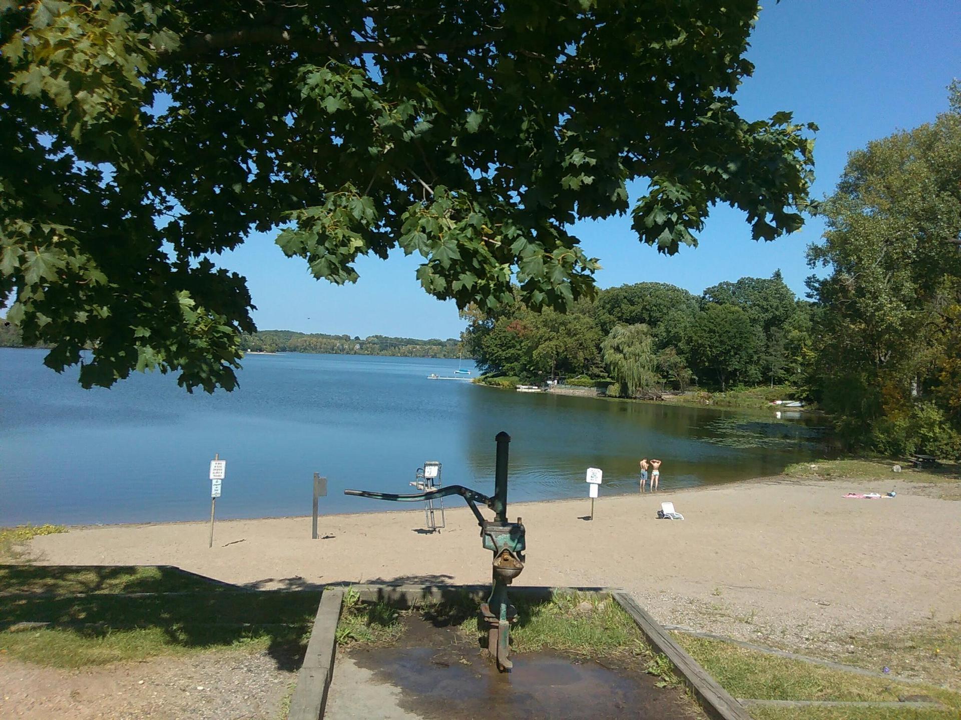 Sandy beach with a hand pump, lake, and people swimming under a clear sky.
