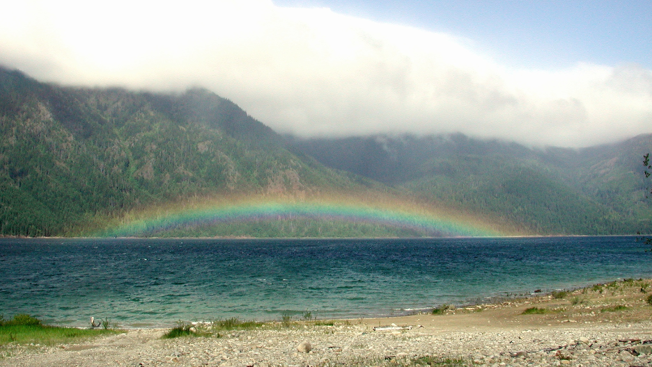 Rainbow over a lake with forested mountains under a cloudy sky.