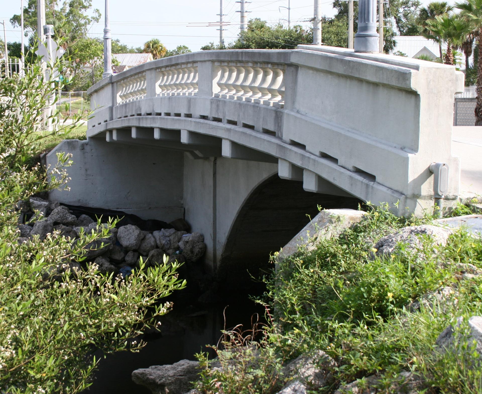 Concrete arched bridge over a small waterway, surrounded by green foliage.