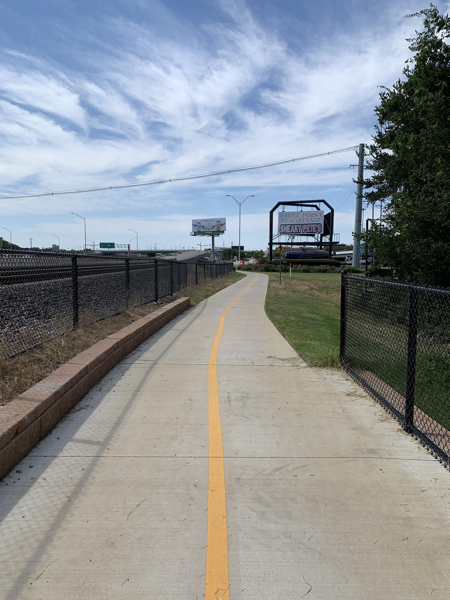 Paved path with yellow line, fences, and distant billboards under cloudy sky.
