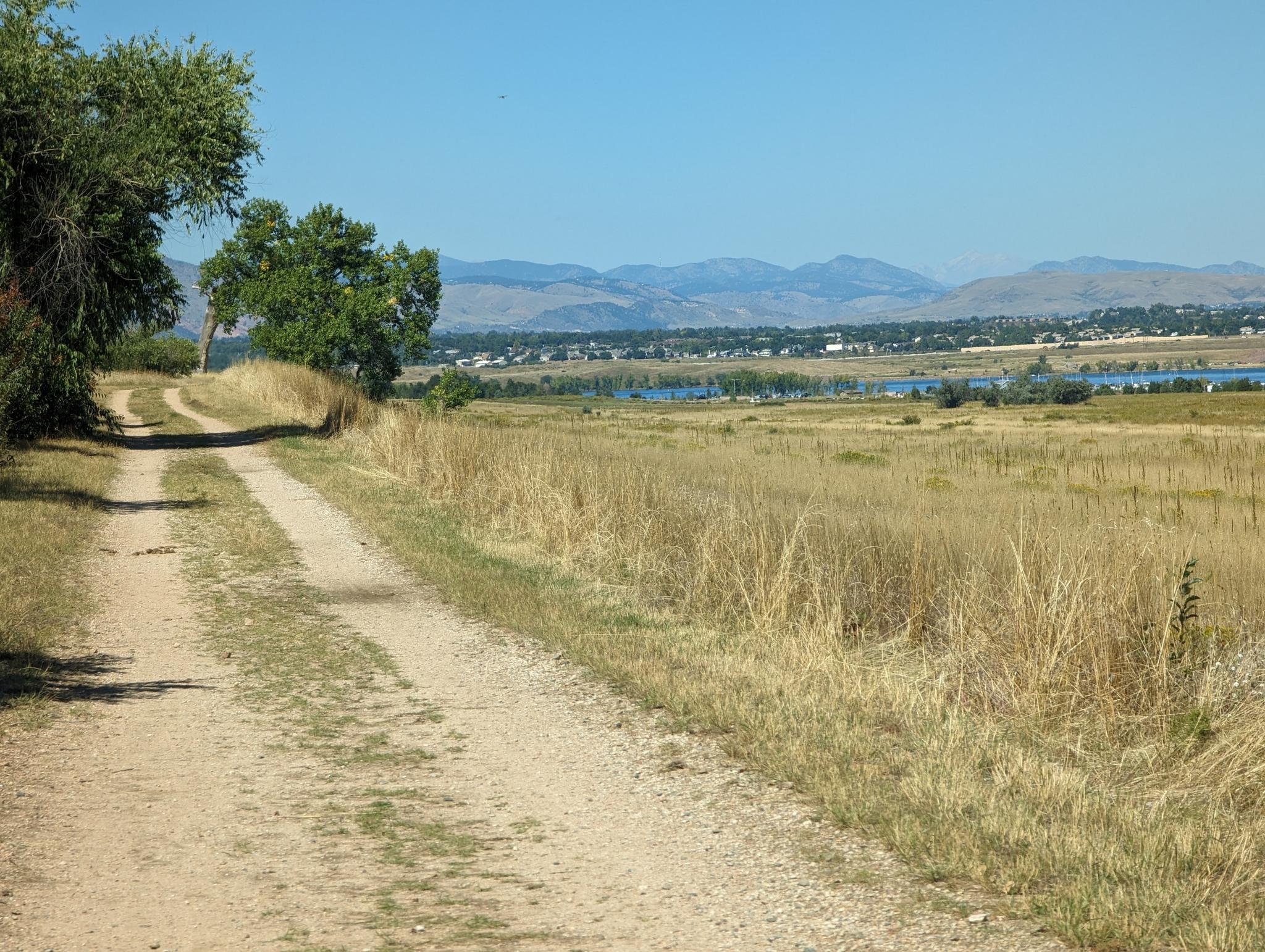 A dirt road winds through dry fields towards a distant lake and mountains.