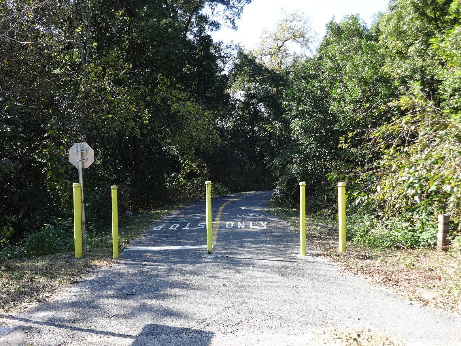 A paved path blocked by yellow bollards, surrounded by dense green trees.