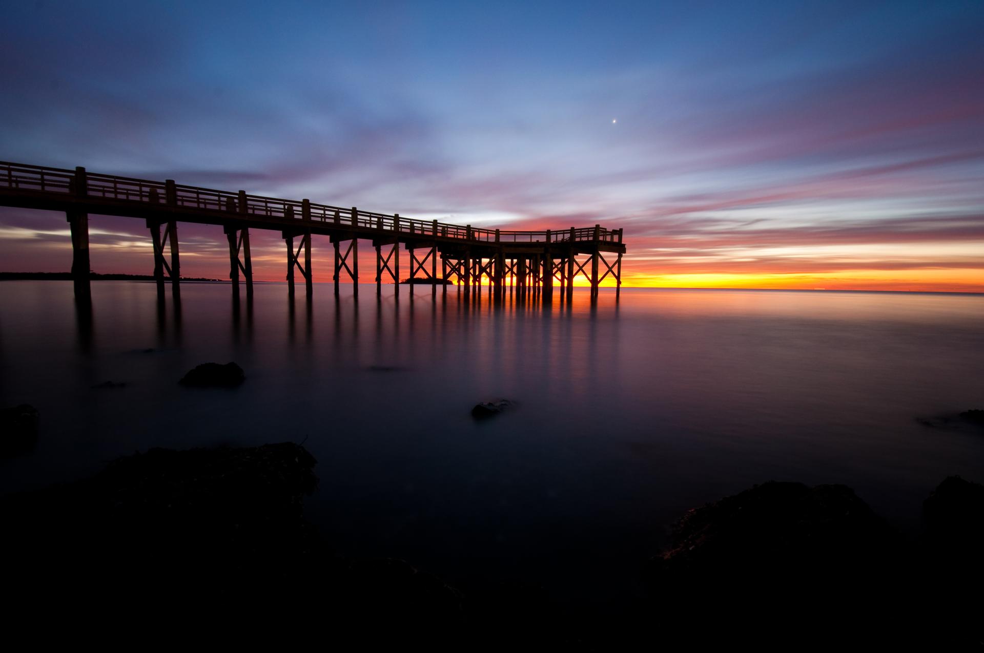 Silhouetted wooden pier extends over water during a vibrant sunset or sunrise.