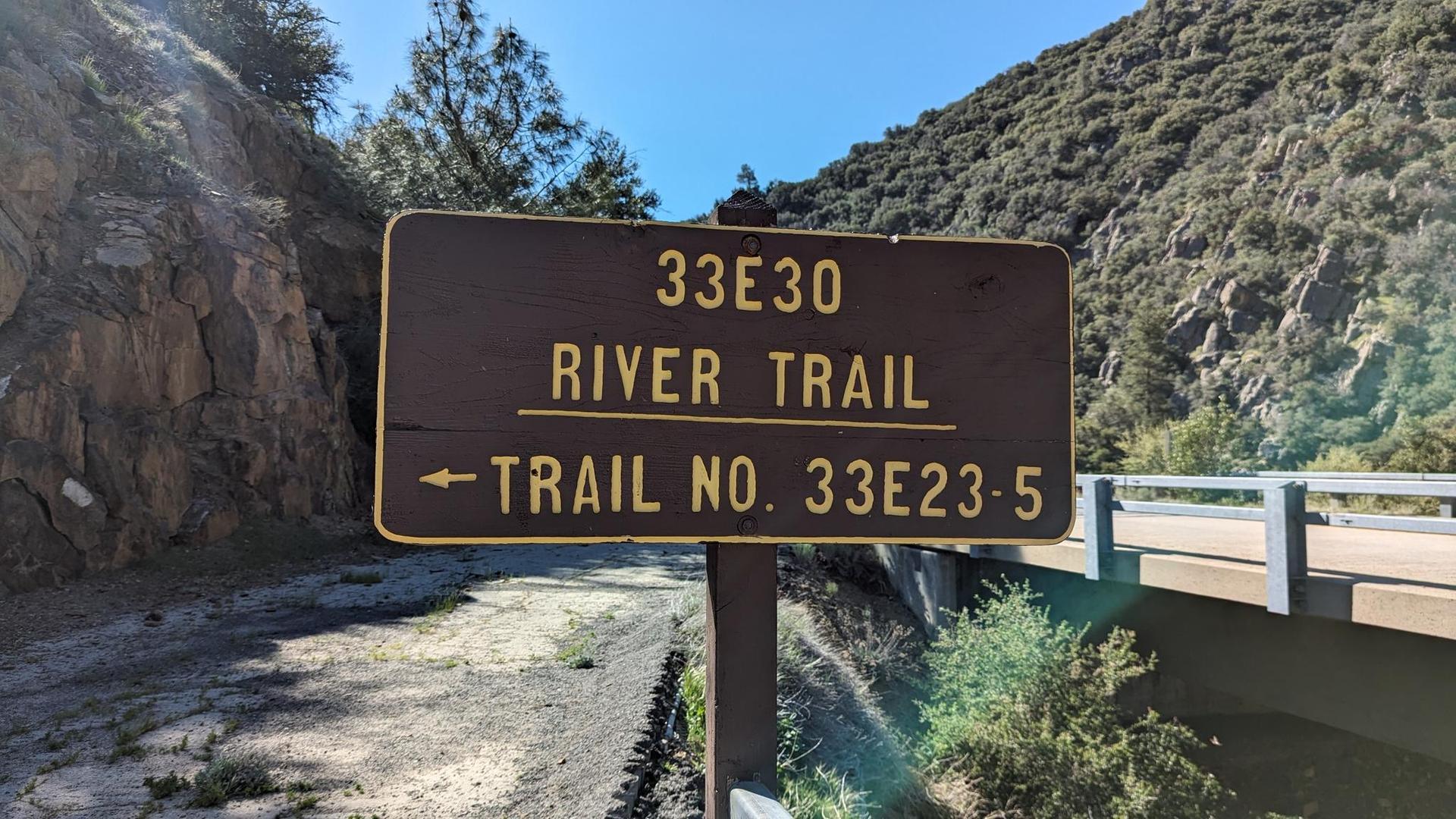 Brown wooden sign reading "RIVER TRAIL" next to a bridge, sunny day.