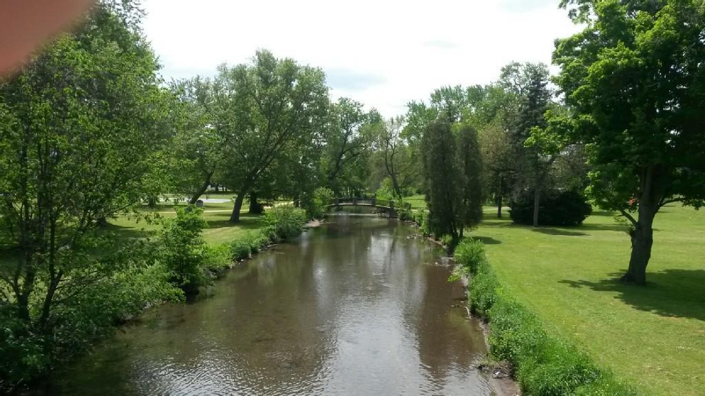 A calm river flows through a lush green park under bright skies.