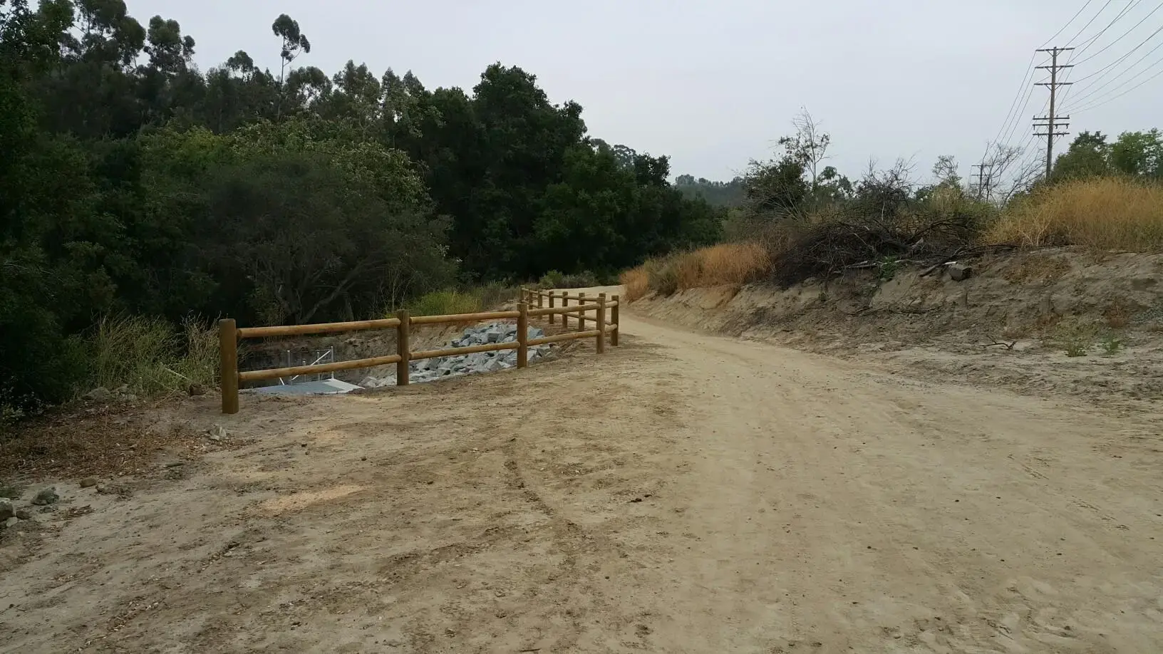 Dirt path, wooden fence, dry creek bed, and dense trees under overcast sky.