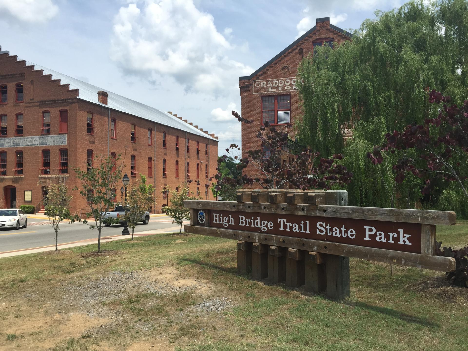 A wooden sign for a trail park in front of old brick buildings, sunny.