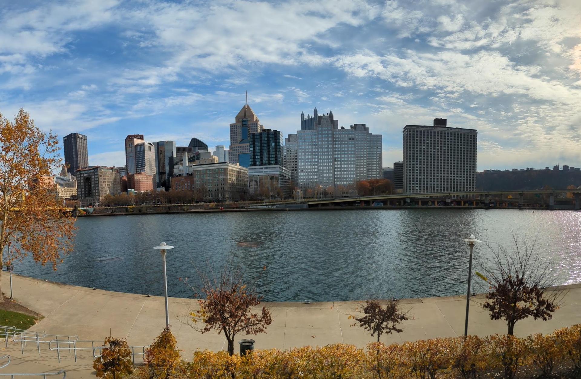 Panoramic city skyline across a wide river with autumn trees under a bright sky.