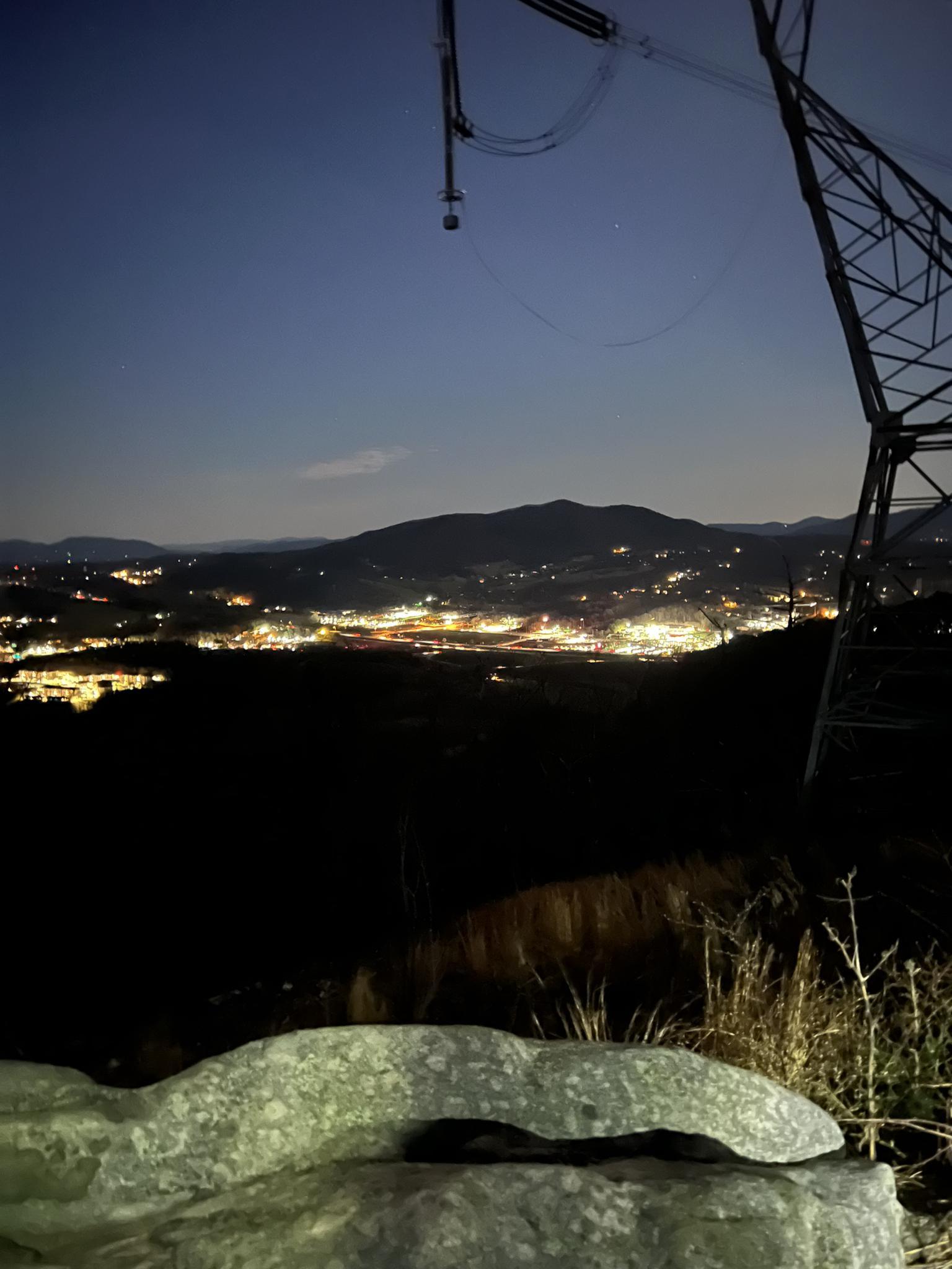 Night view of a city's lights from a mountain, power tower visible.
