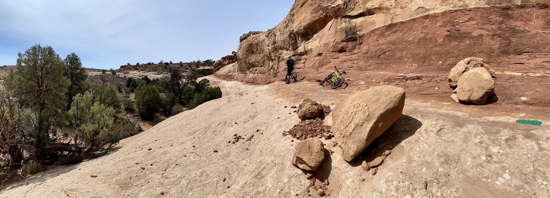 Person with mountain bike on a narrow rocky trail beside a large cliff.