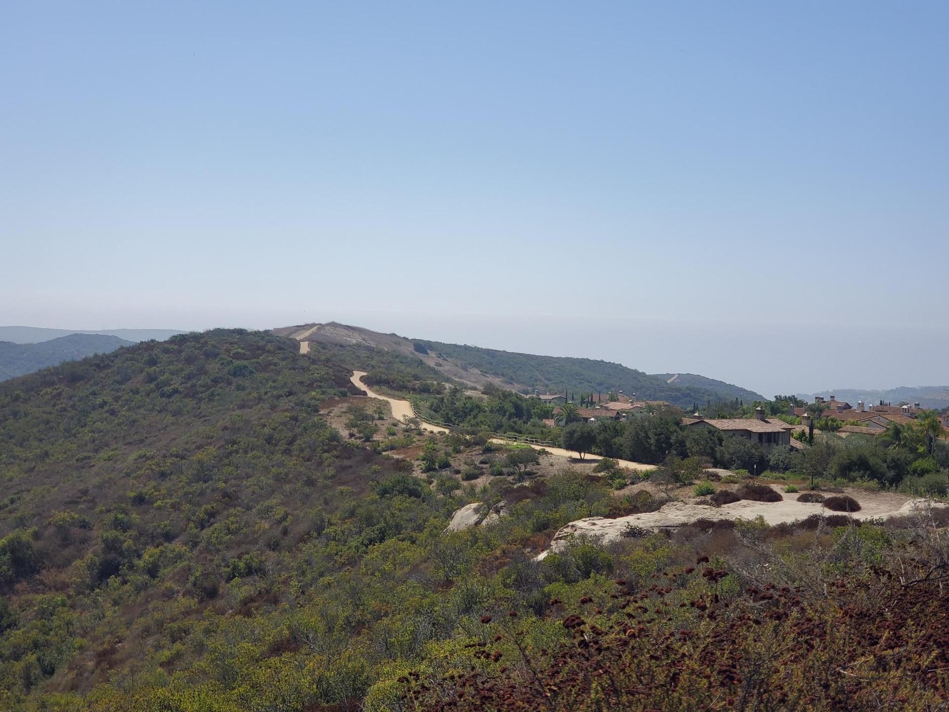 A dirt road winds through green hills with distant houses under a bright blue sky.
