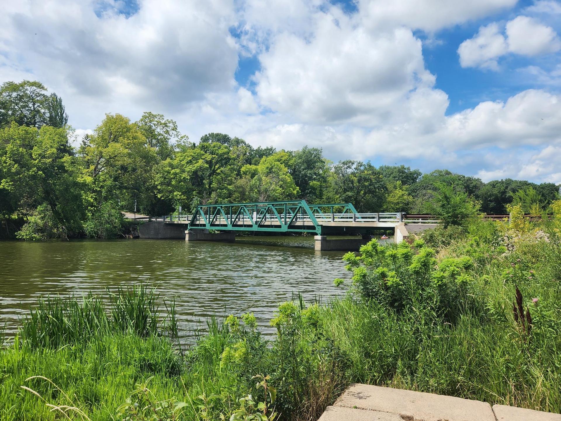 Green truss bridge over a river, surrounded by lush green foliage.