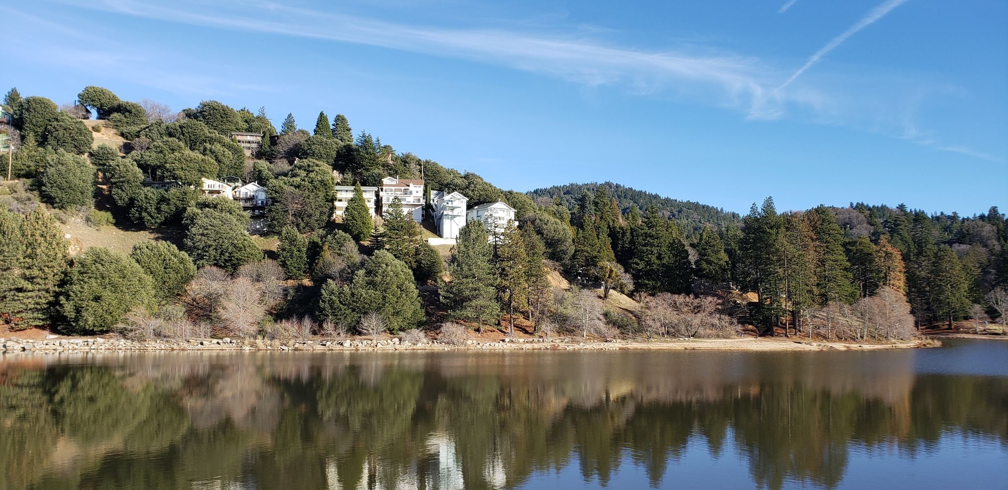 Calm lake reflecting white buildings and green trees on a sunny hillside. - Lake Gregory loop from Lake Gregory