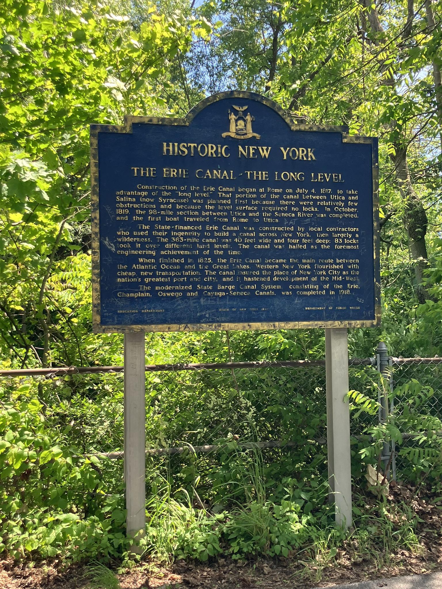 A blue historic sign with gold text, surrounded by green foliage on a sunny day.