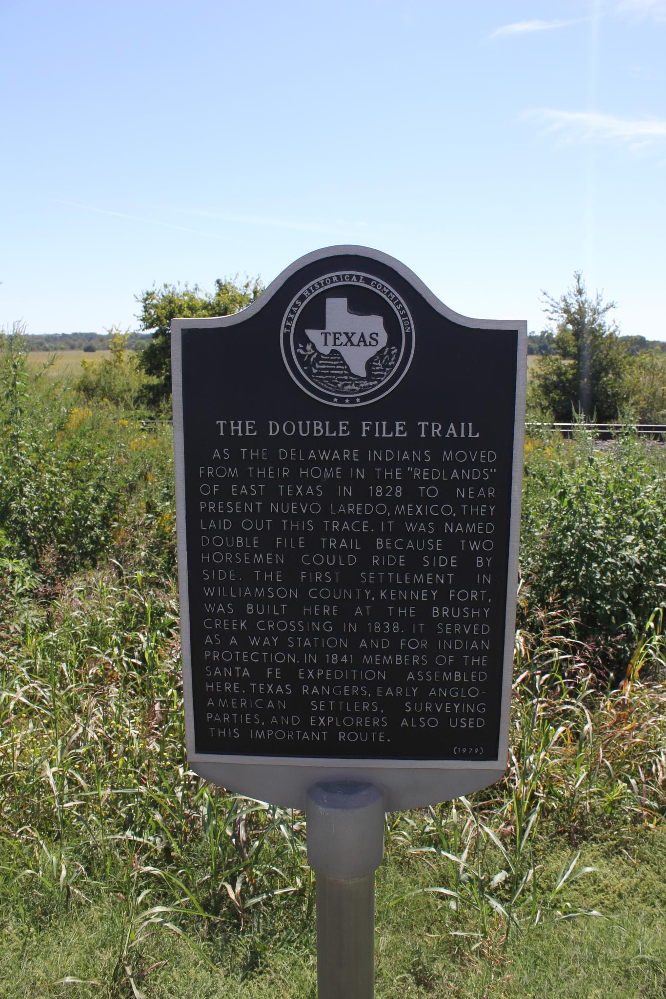 A black historical marker stands in a sunny, grassy field with distant trees.