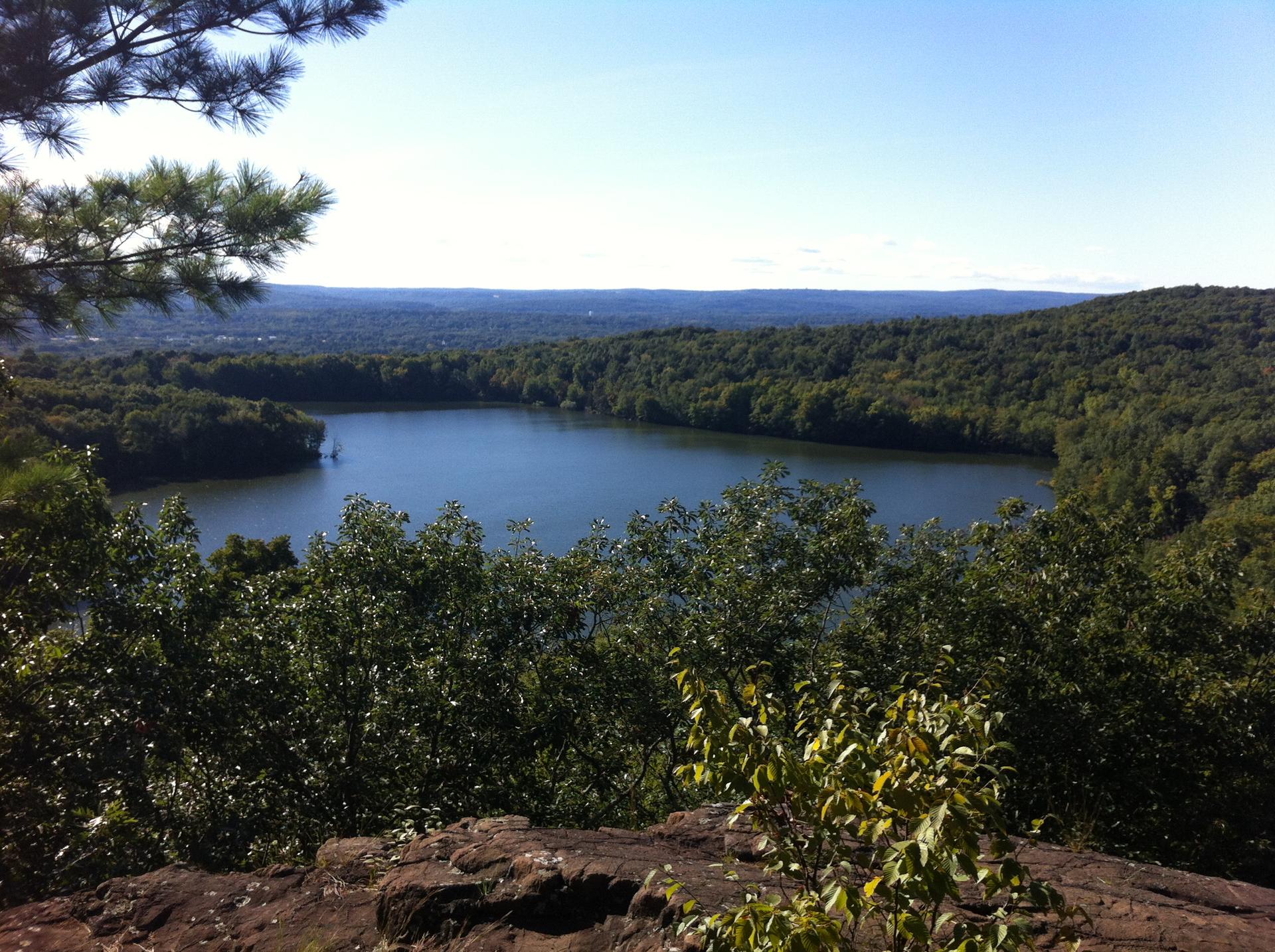 A scenic overlook shows a calm lake surrounded by dense green forests under a clear sky.