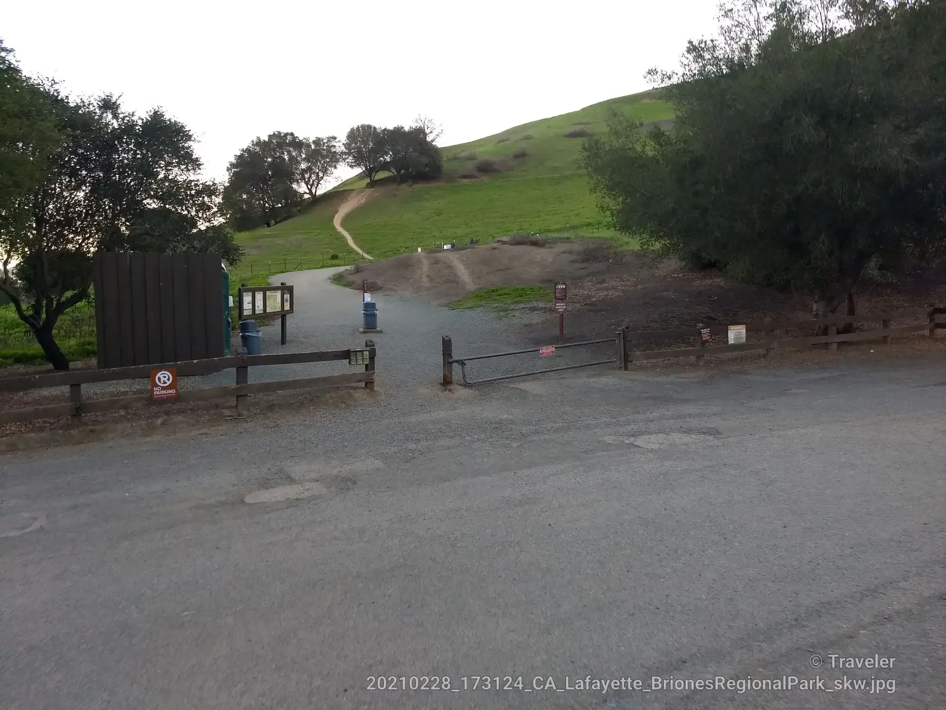 Trailhead entrance with a metal gate, signs, and a path leading up a green hill.