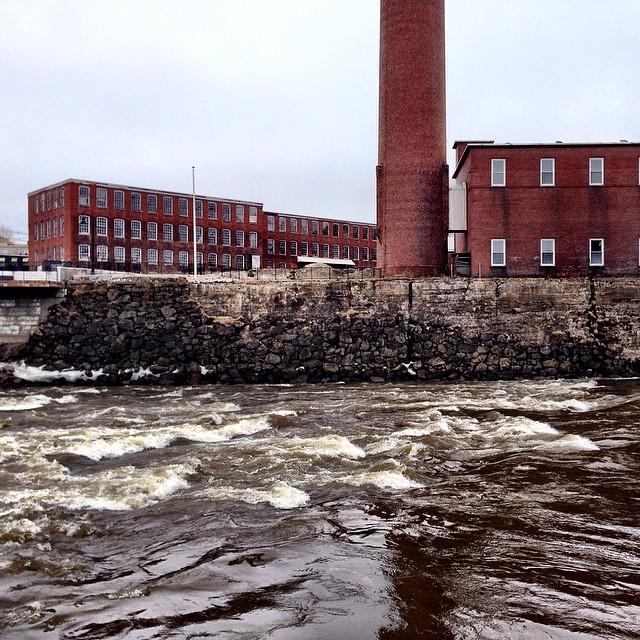 Turbulent river flowing past large red brick industrial buildings under an overcast sky.