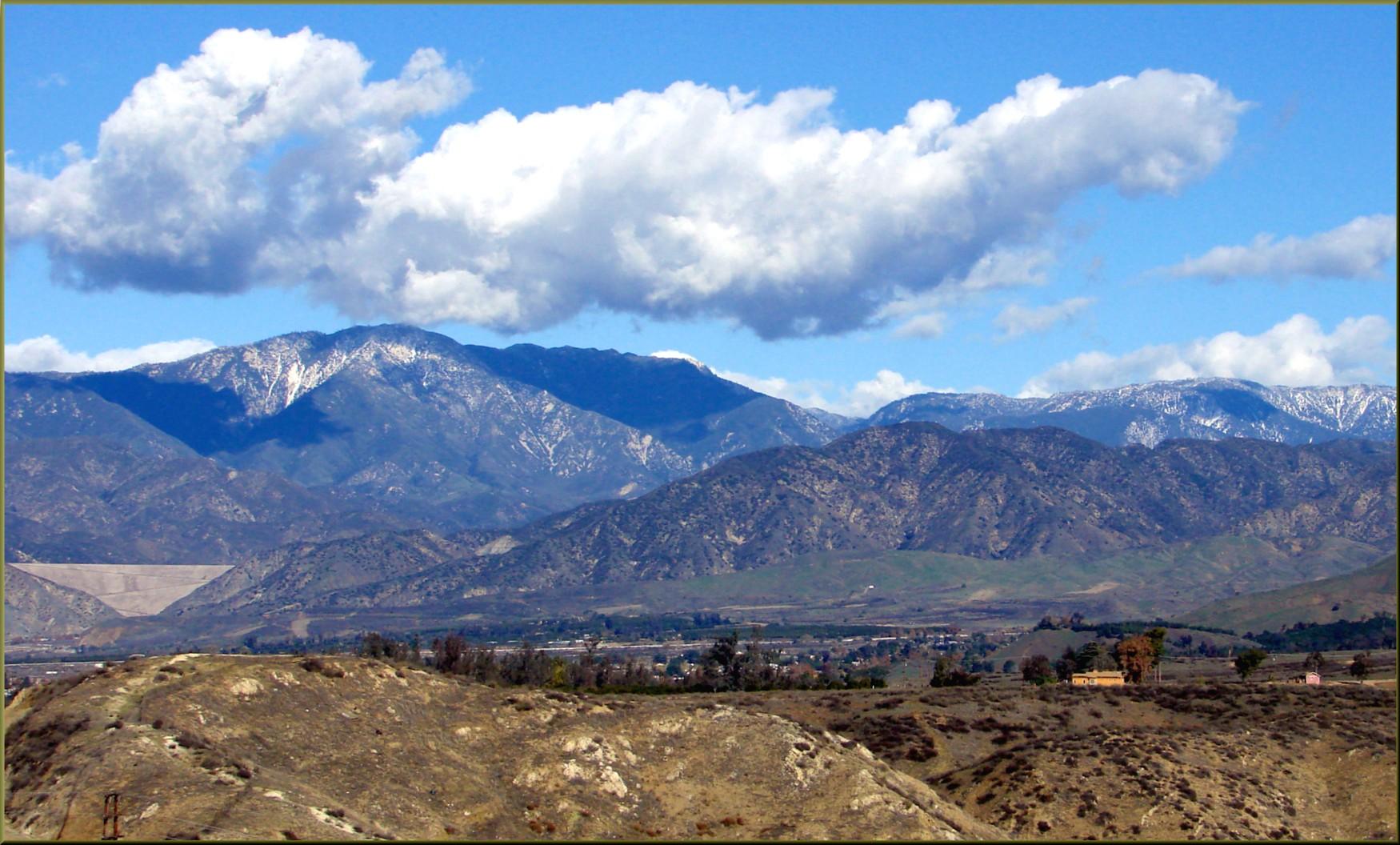 Snow-capped mountains under large white clouds with dry, rolling hills below.