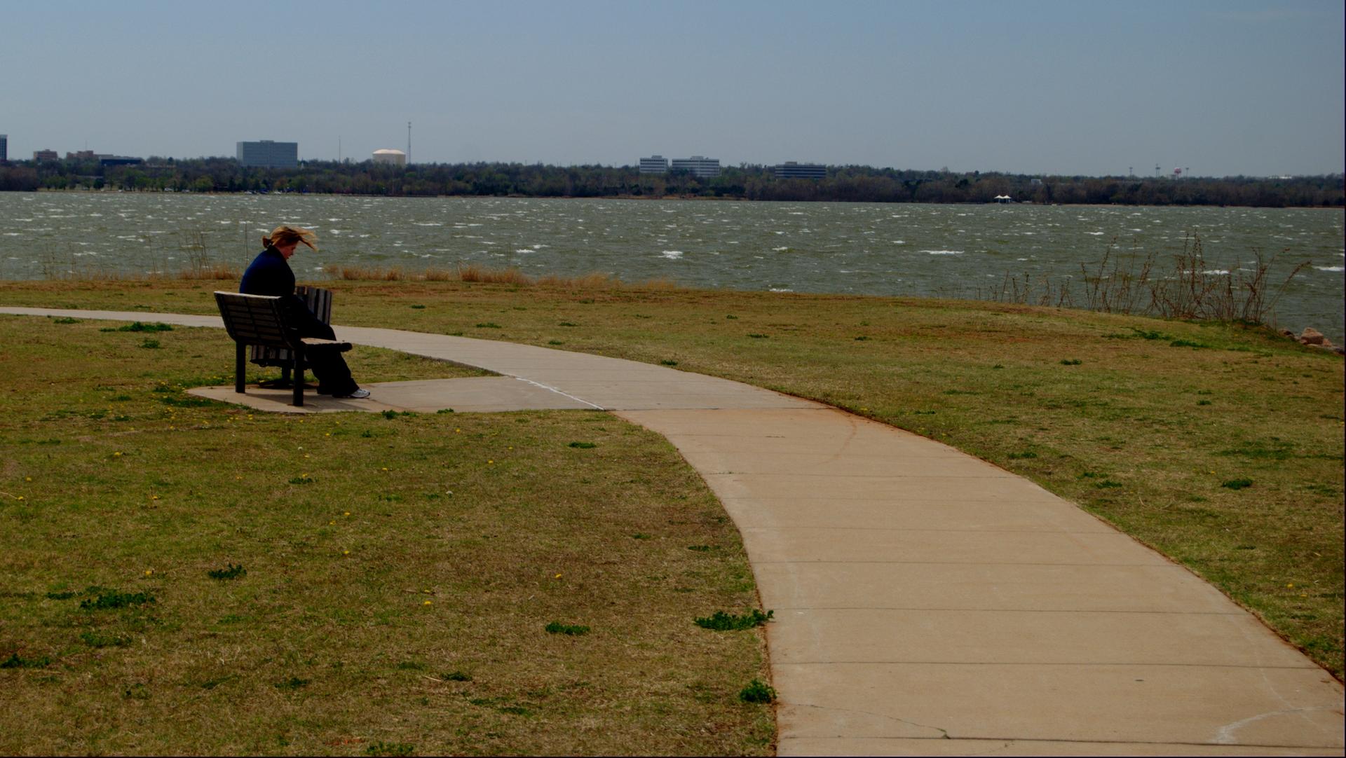 Person sits on a bench beside a paved path overlooking wavy water.