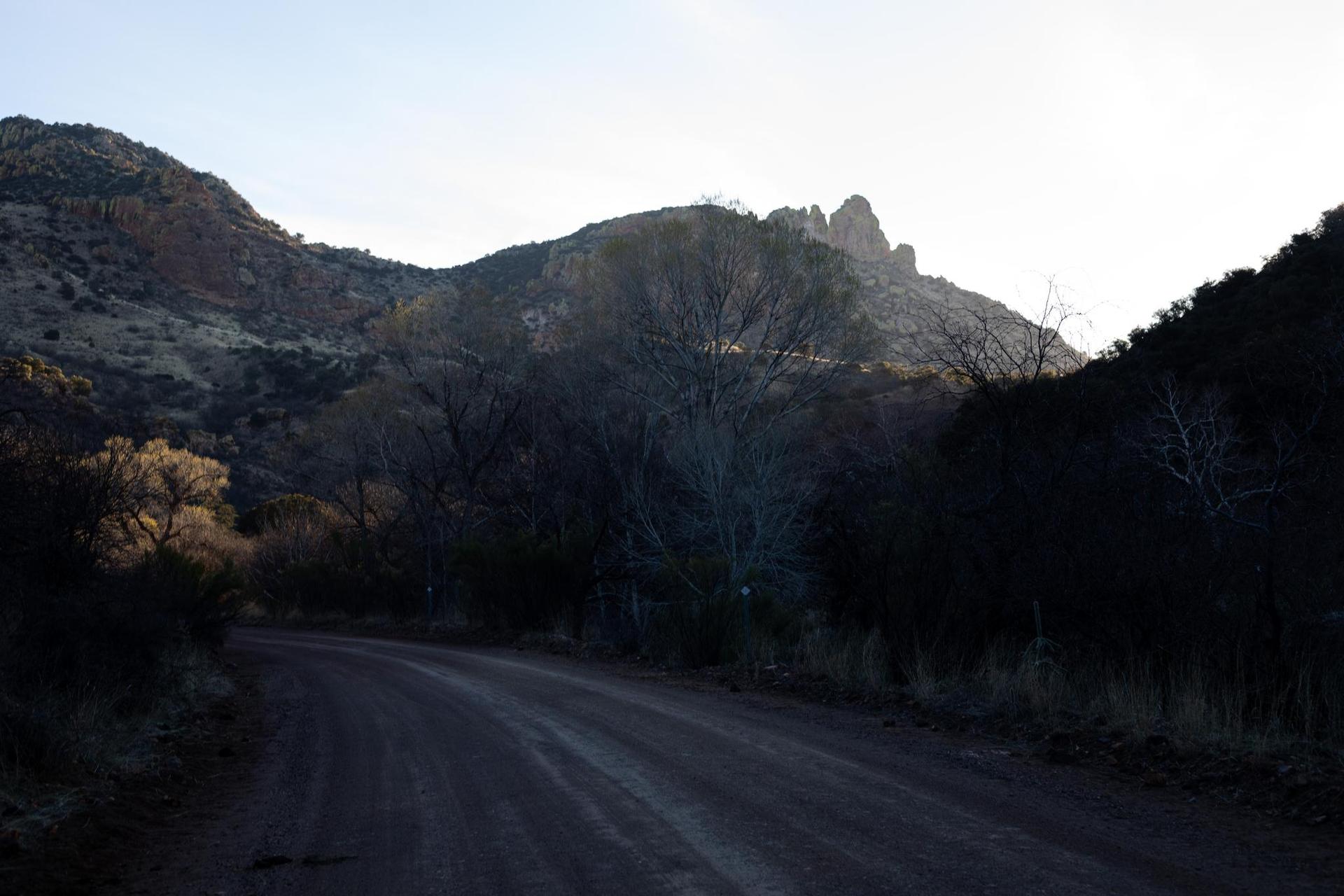 A dirt road winds through a dark valley with sunlit mountains in the distance.