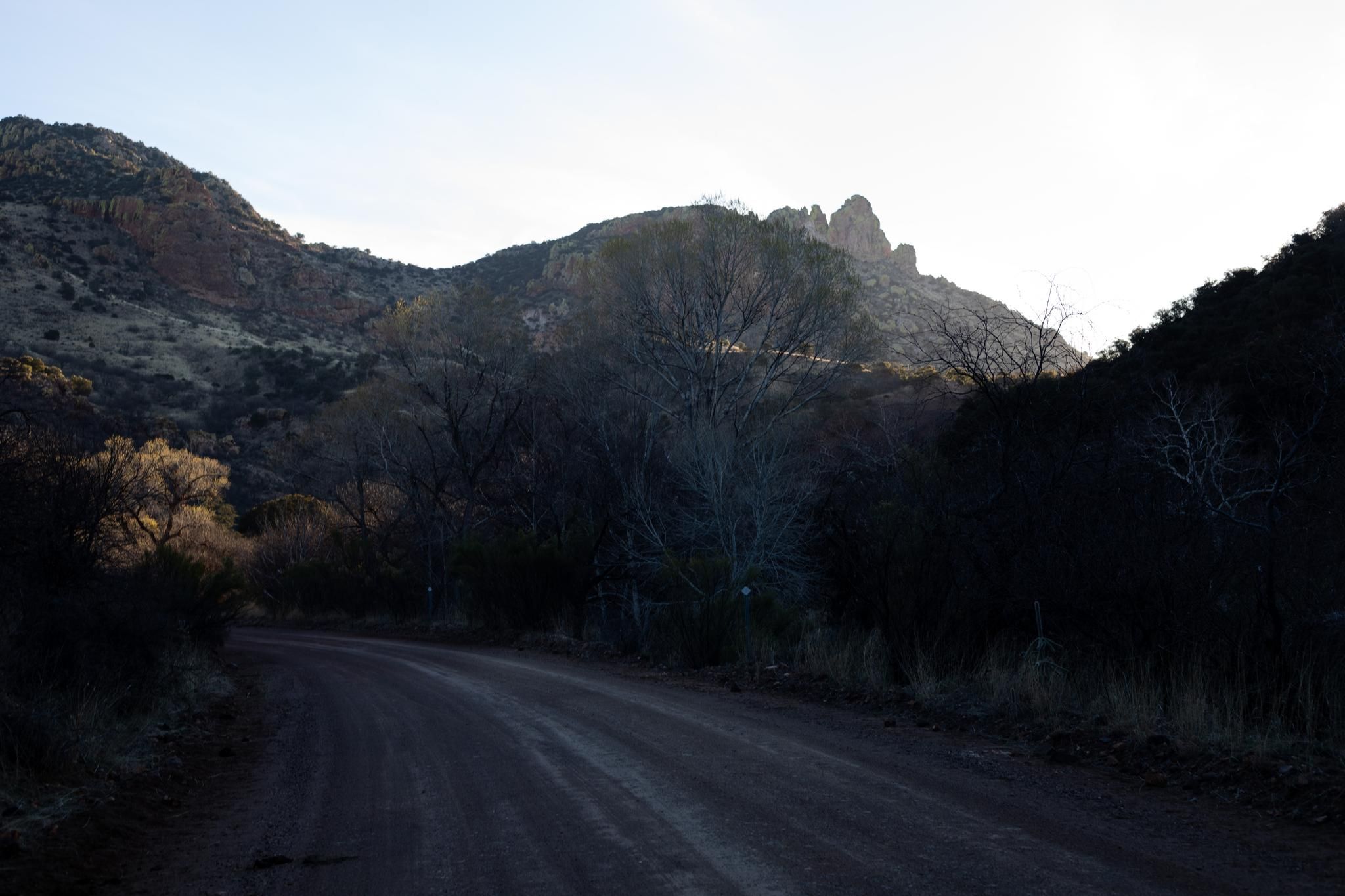 A dirt road winds through a dark valley with sunlit mountains in the distance. - Apache Road (Gravel) loop from Patagonia