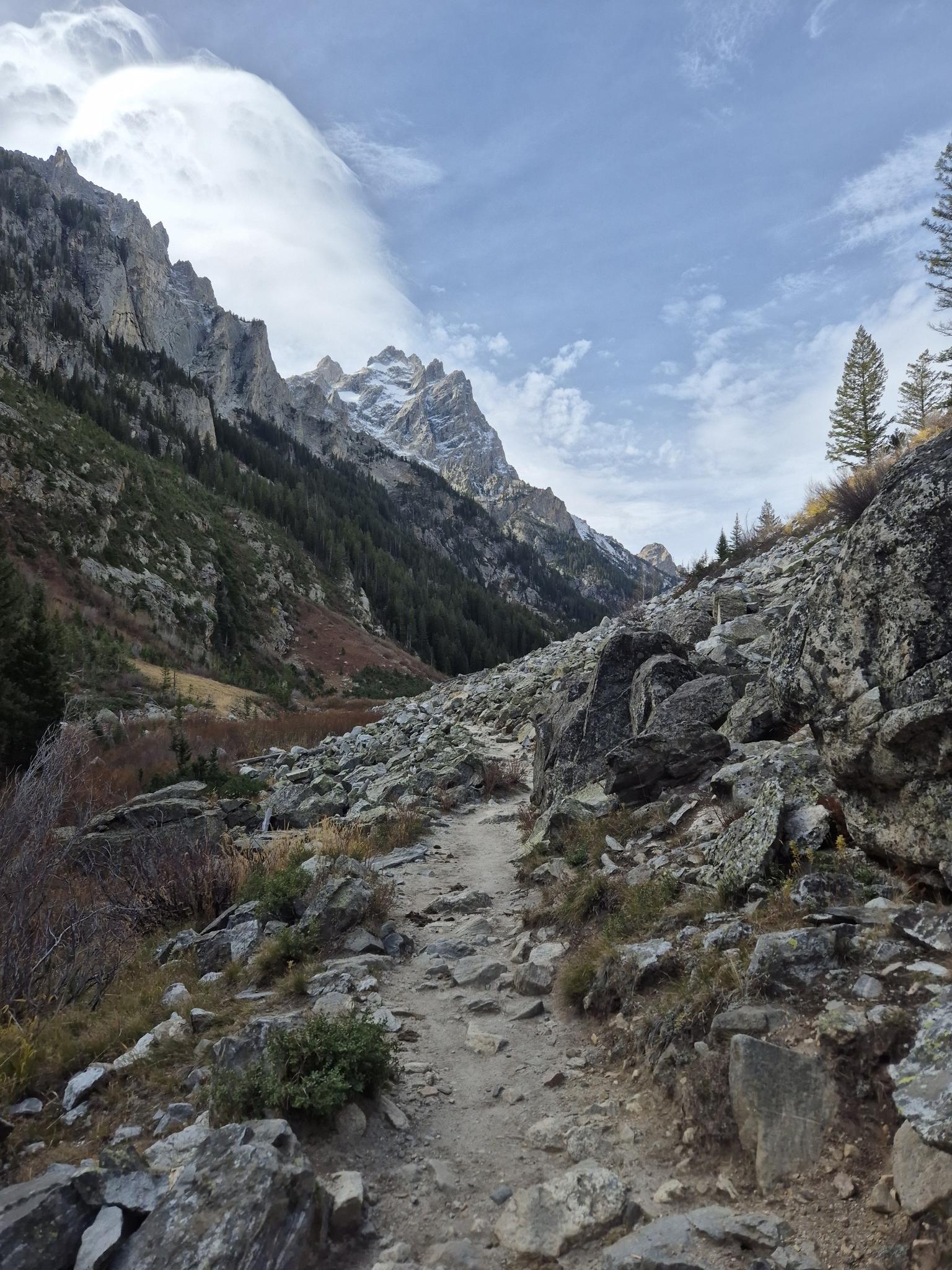 A rocky trail winds through a mountain valley with steep slopes and distant peaks.
