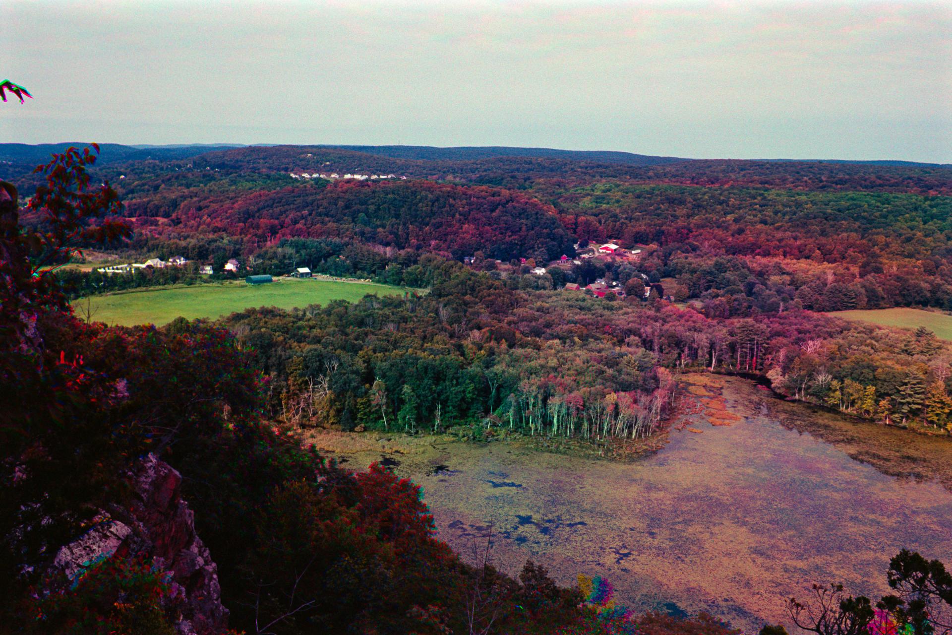 Overlook of a lake surrounded by autumn-colored forest and fields under a hazy sky.
