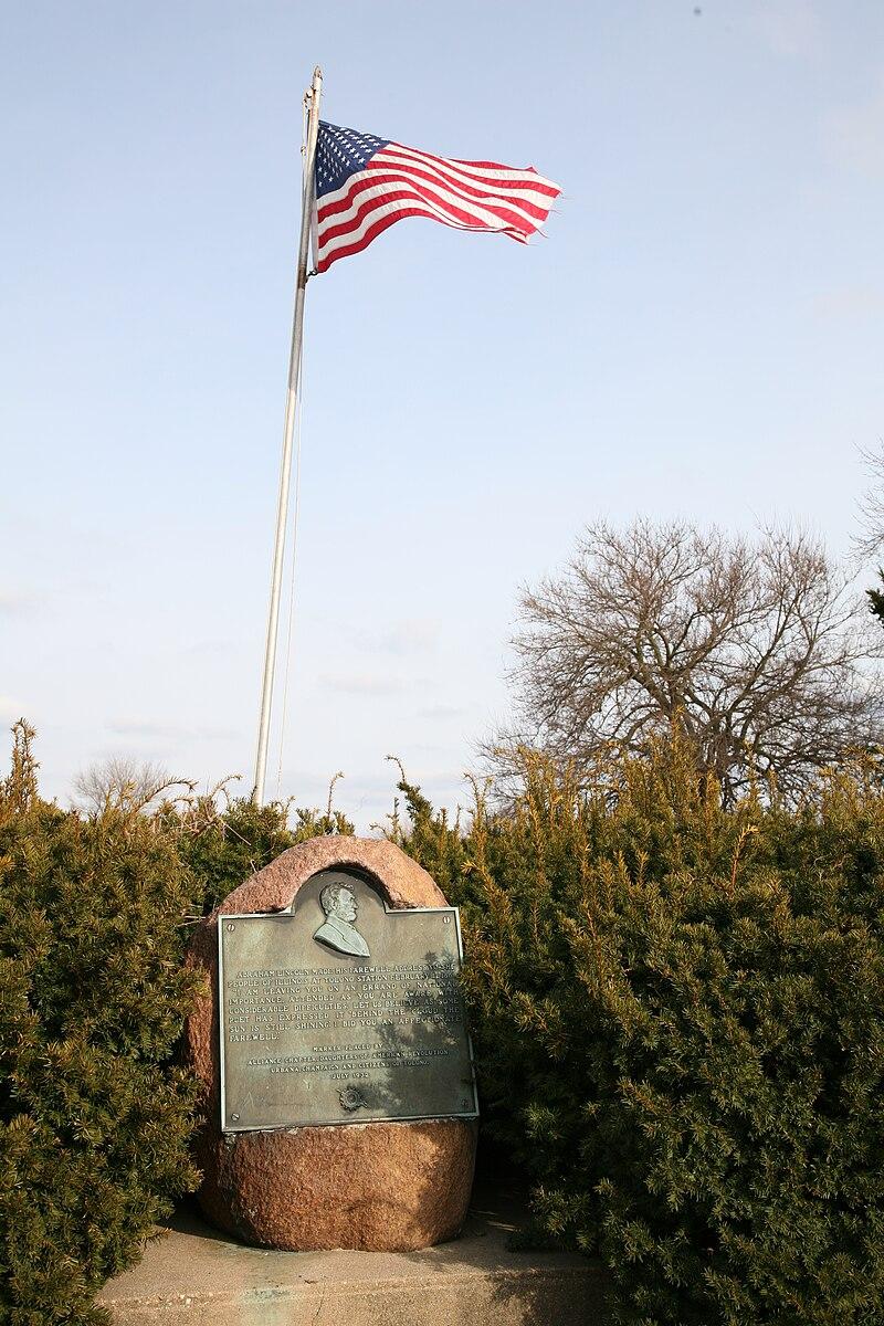 American flag flying above a historical marker surrounded by green bushes.