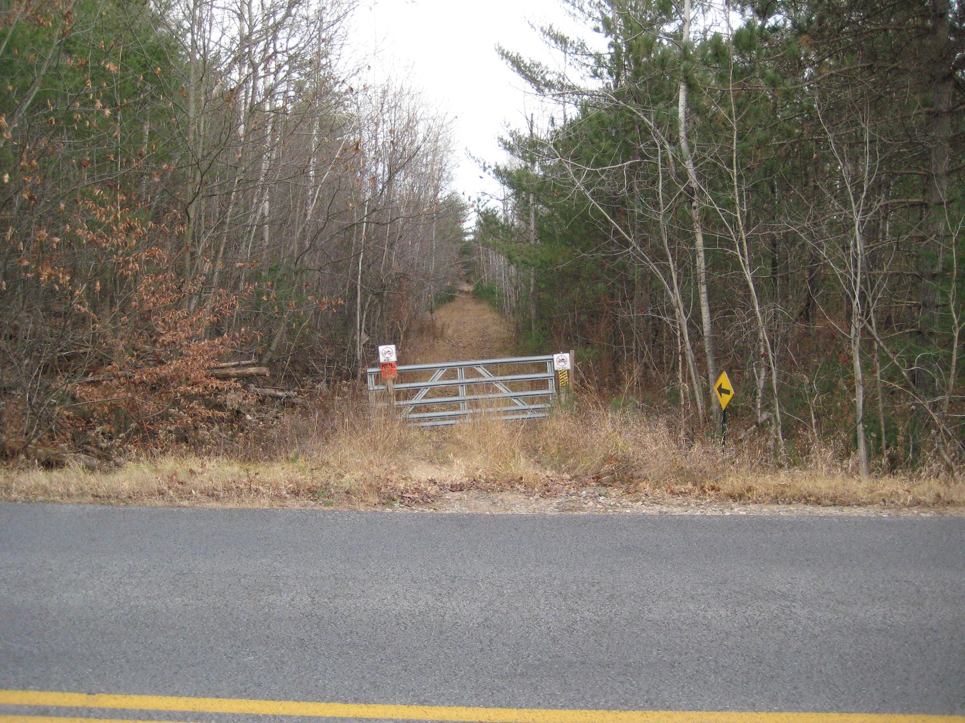 A metal gate blocks a dirt path into a forest under overcast skies.