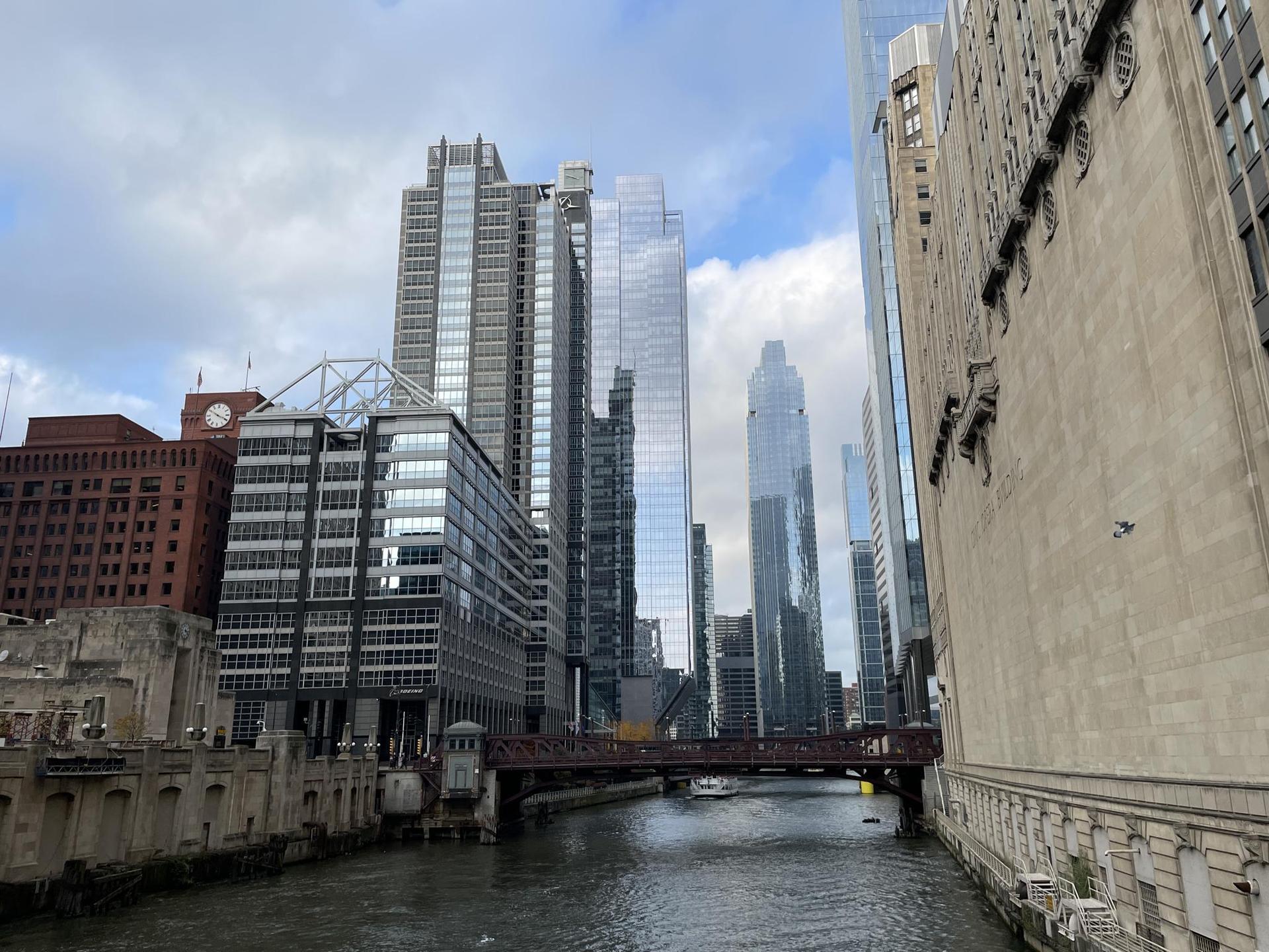 A river flows beneath a bridge, surrounded by diverse city architecture under clouds.