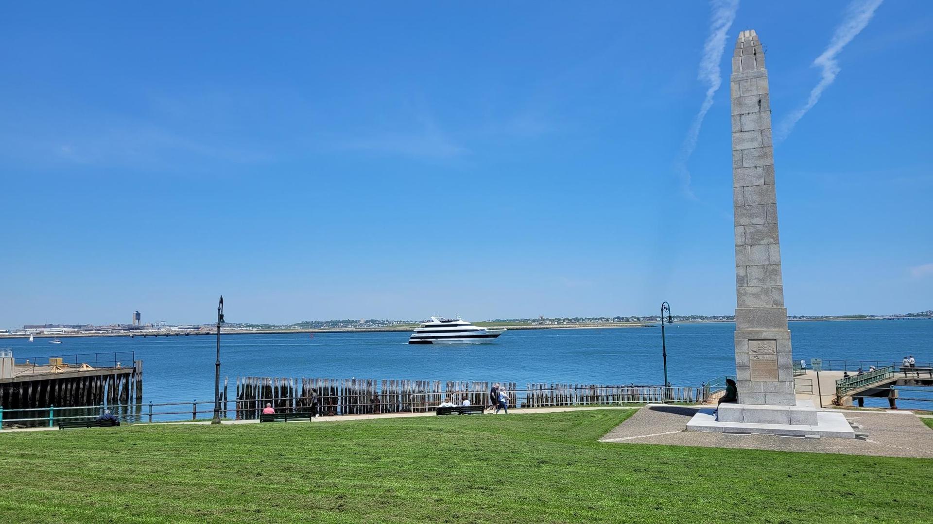 Tall stone obelisk by a bay with a boat, green grass, under bright sun.