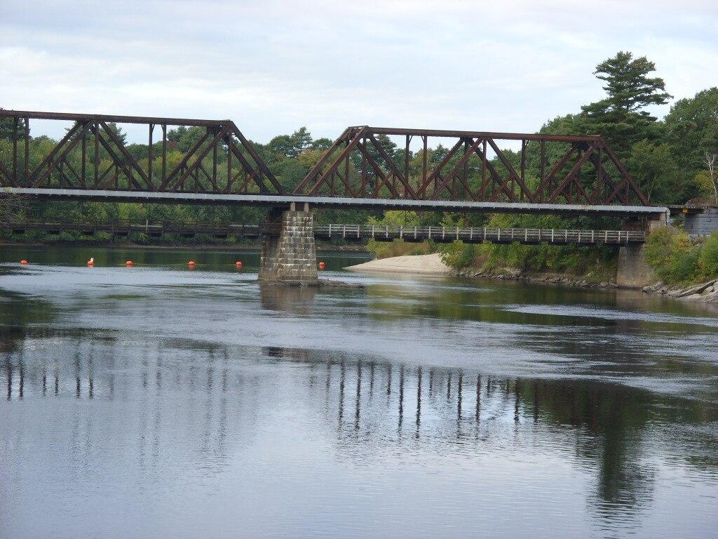 A long metal truss bridge spans a calm river under an overcast sky.
