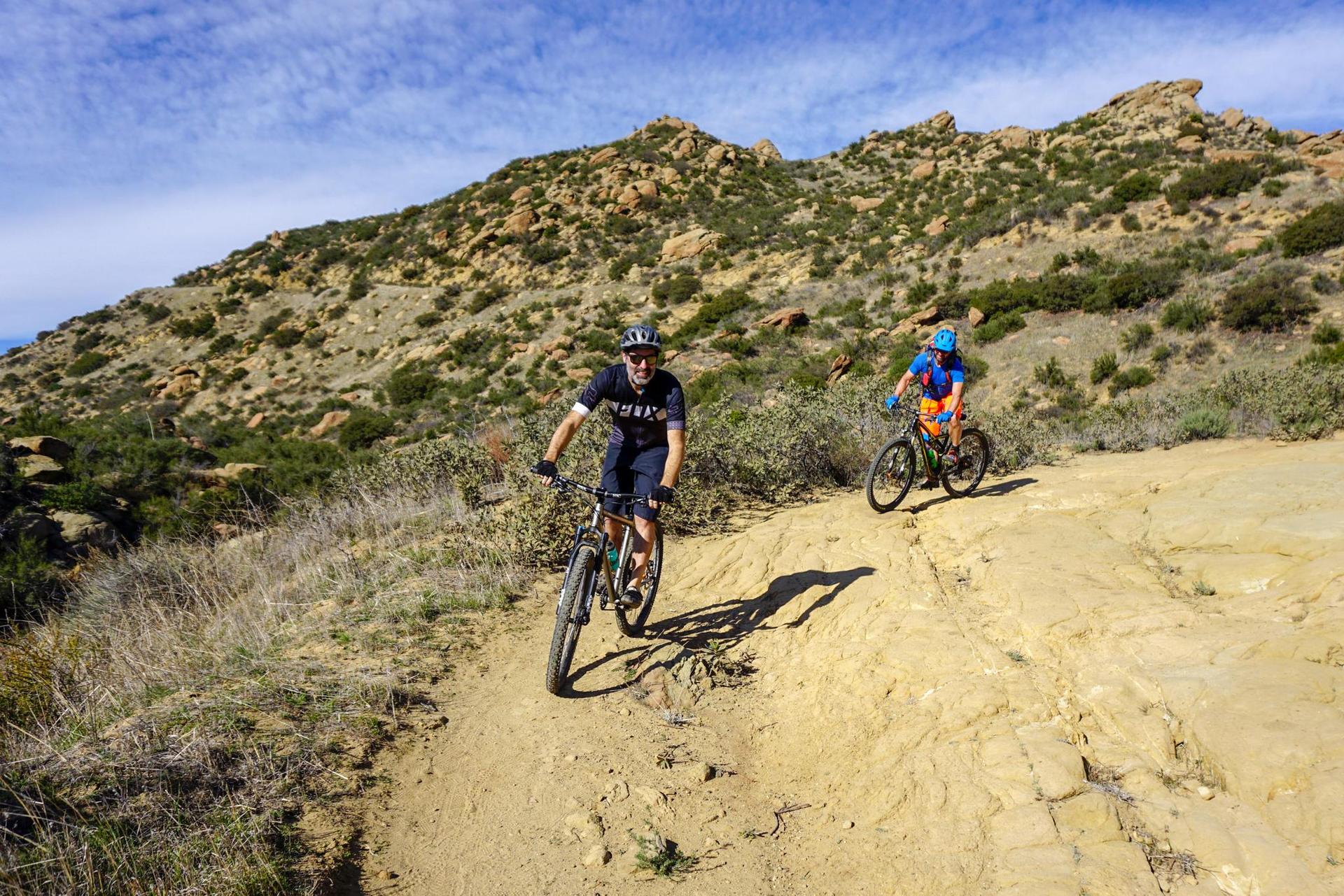 Two mountain bikers ride a sunny, rocky dirt trail through dry hills.