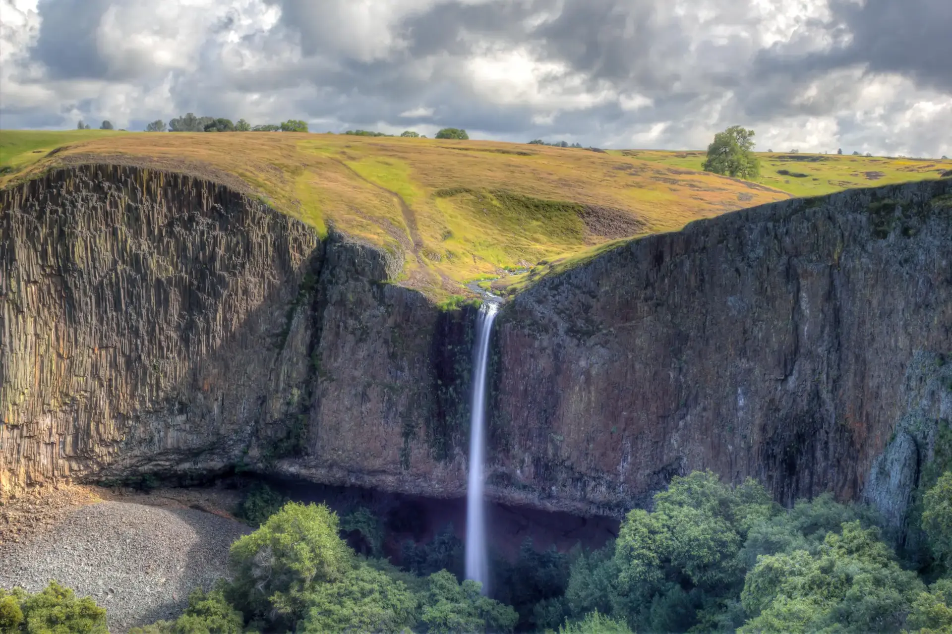 Tall waterfall plunges over dark cliffs into a lush green canyon under cloudy skies.