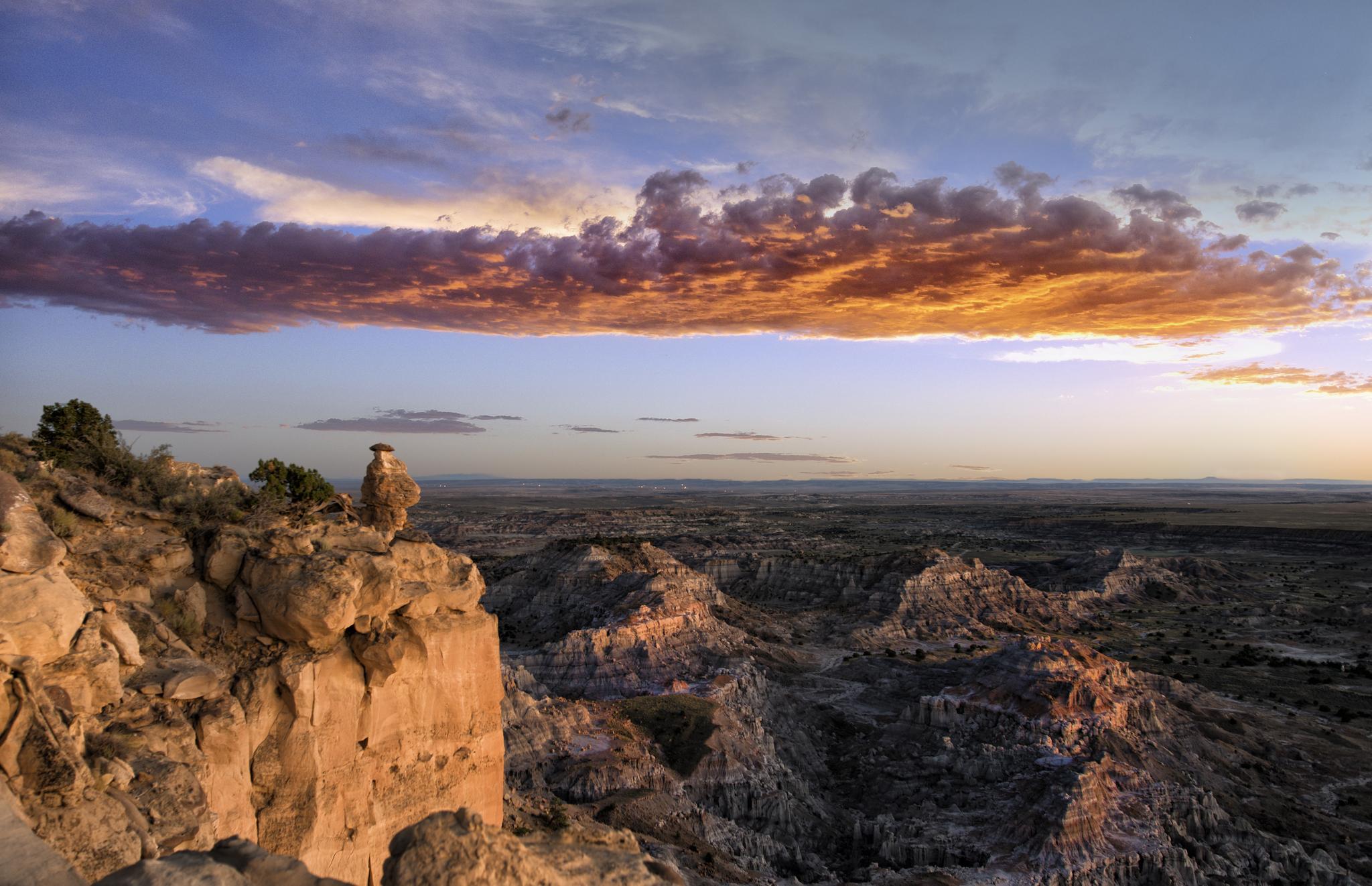 Bird on sunlit cliff overlooking vast rocky landscape under colorful sunset clouds.