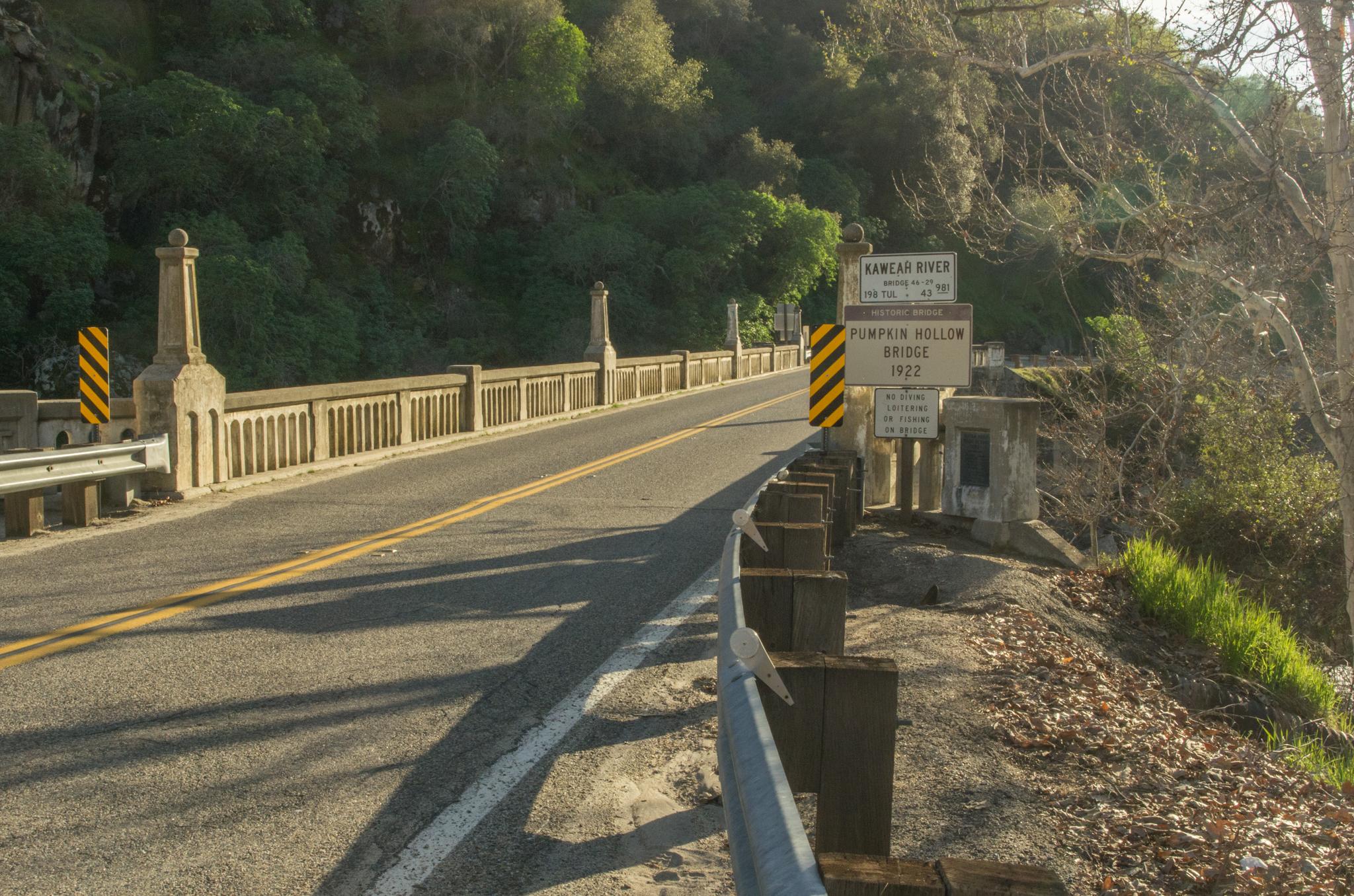 A concrete bridge with a road, sunny, forested hill in the background.