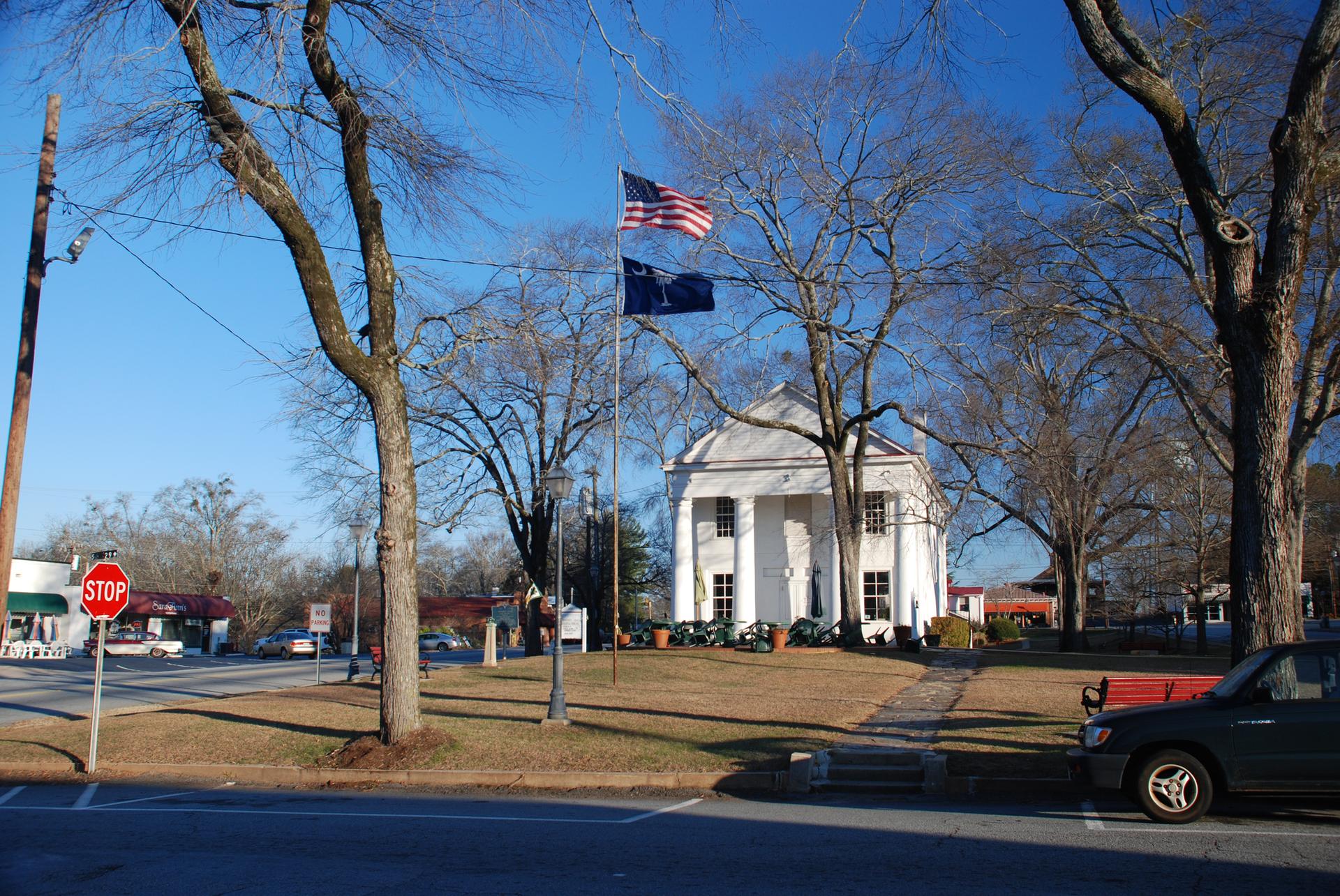 A white building with columns, flags, bare trees, and a stop sign on a sunny day.