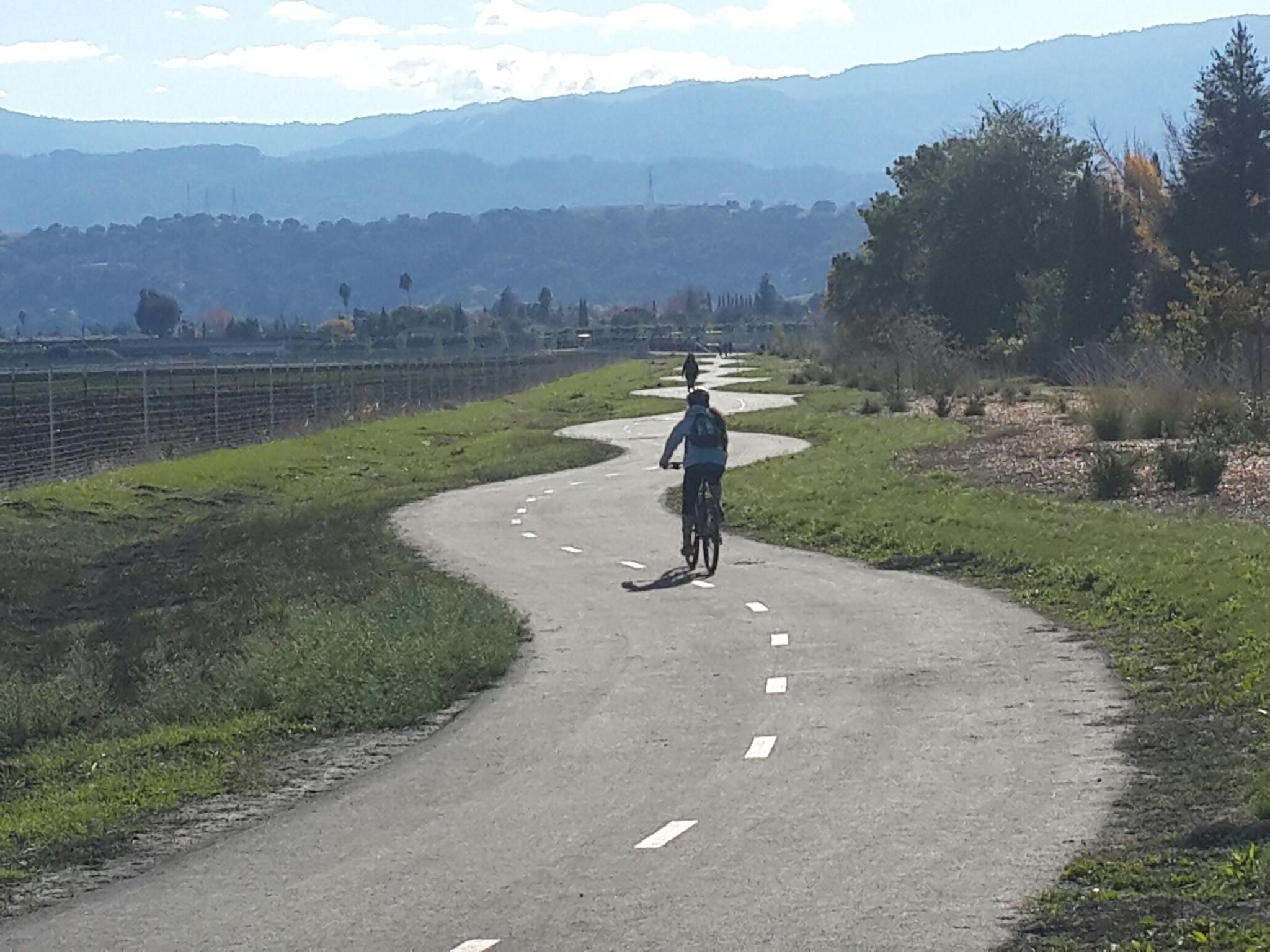 Person cycling on a winding paved path with grass and distant hills. - Edenvale loop from Santa Teresa County Park
