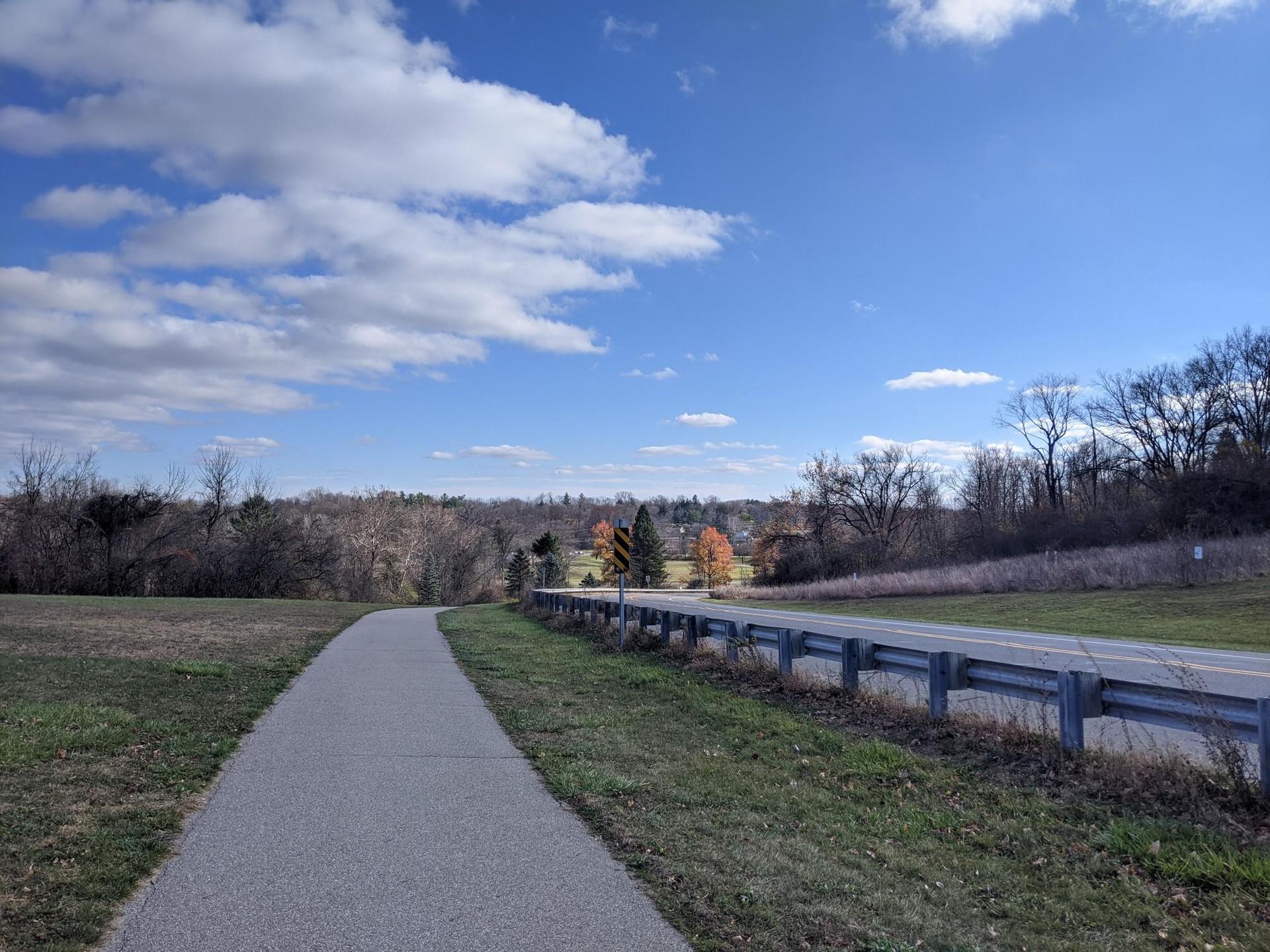 Paved path next to a road with a guardrail under a bright, cloudy sky.