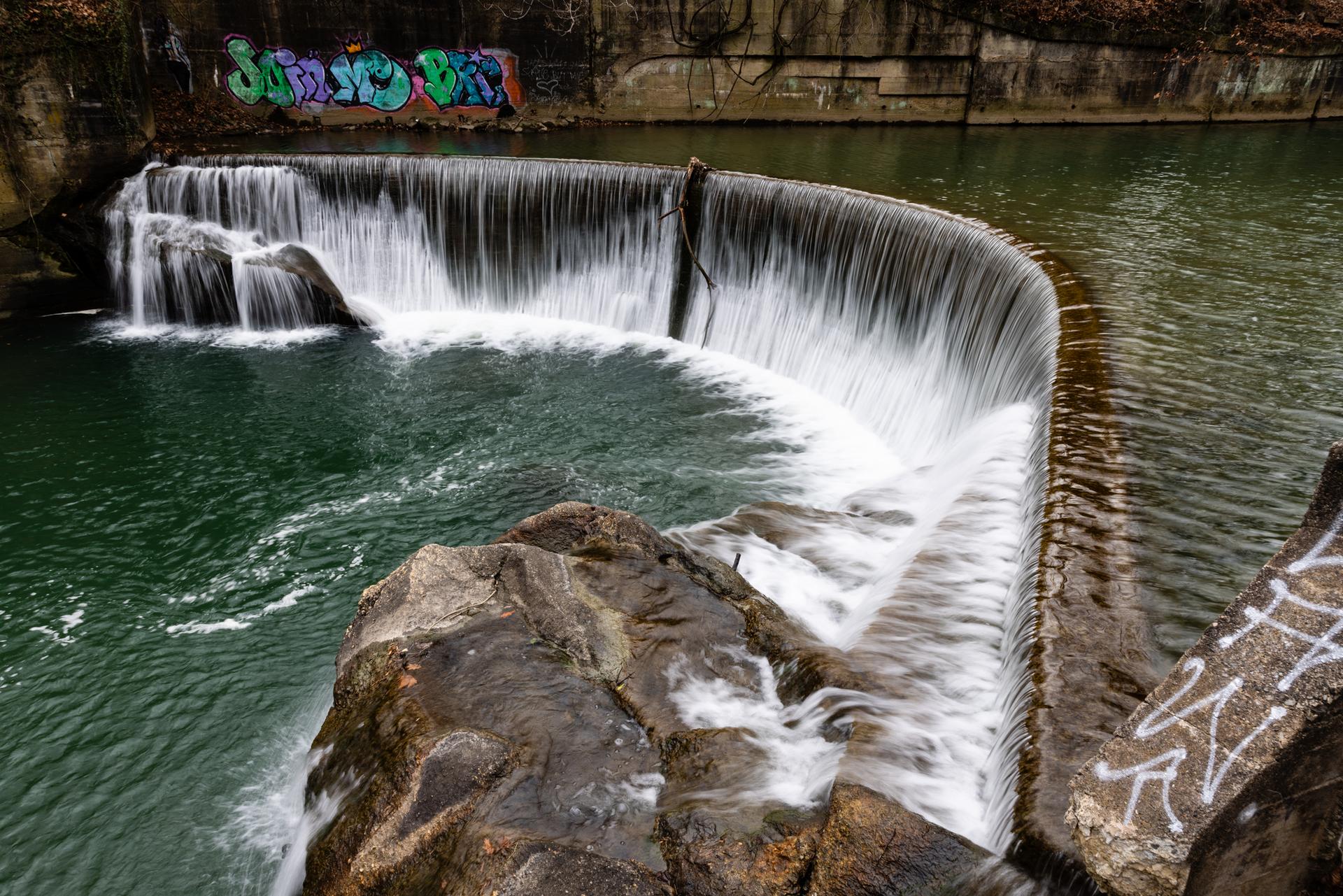 Water cascades over a curved dam into a dark pool, with graffiti visible.