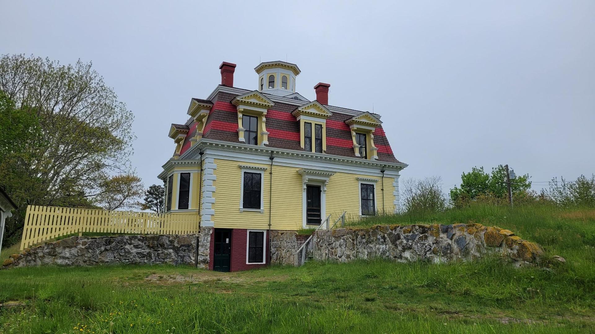 A large yellow house with a red roof on a grassy hill, overcast day.