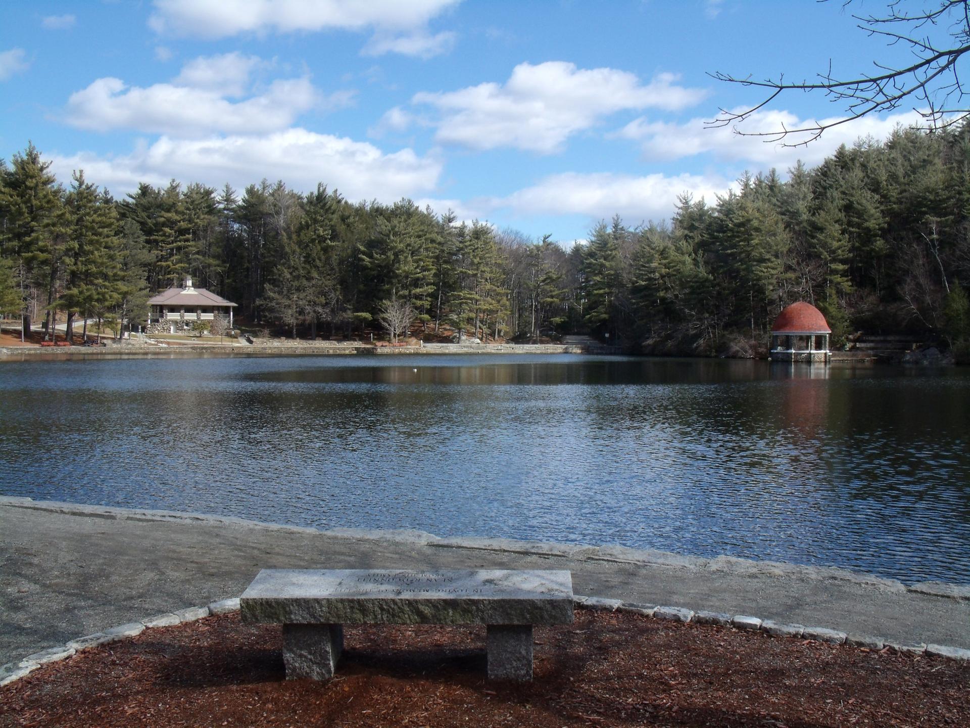 Lake with forested shores, a building, a gazebo, and a stone bench on a sunny day.