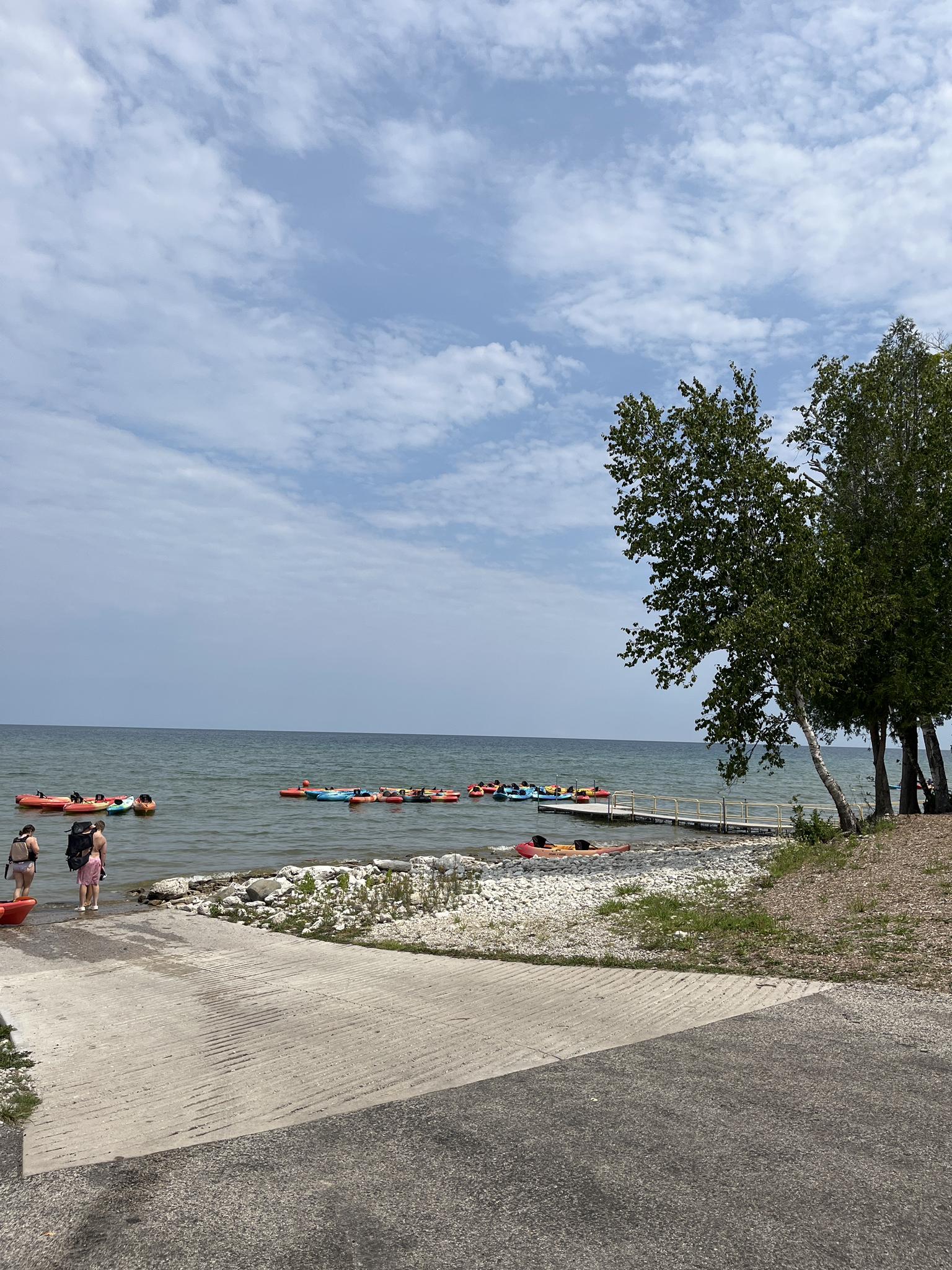 People and inflatable tubes in calm water near a boat ramp under a cloudy sky.