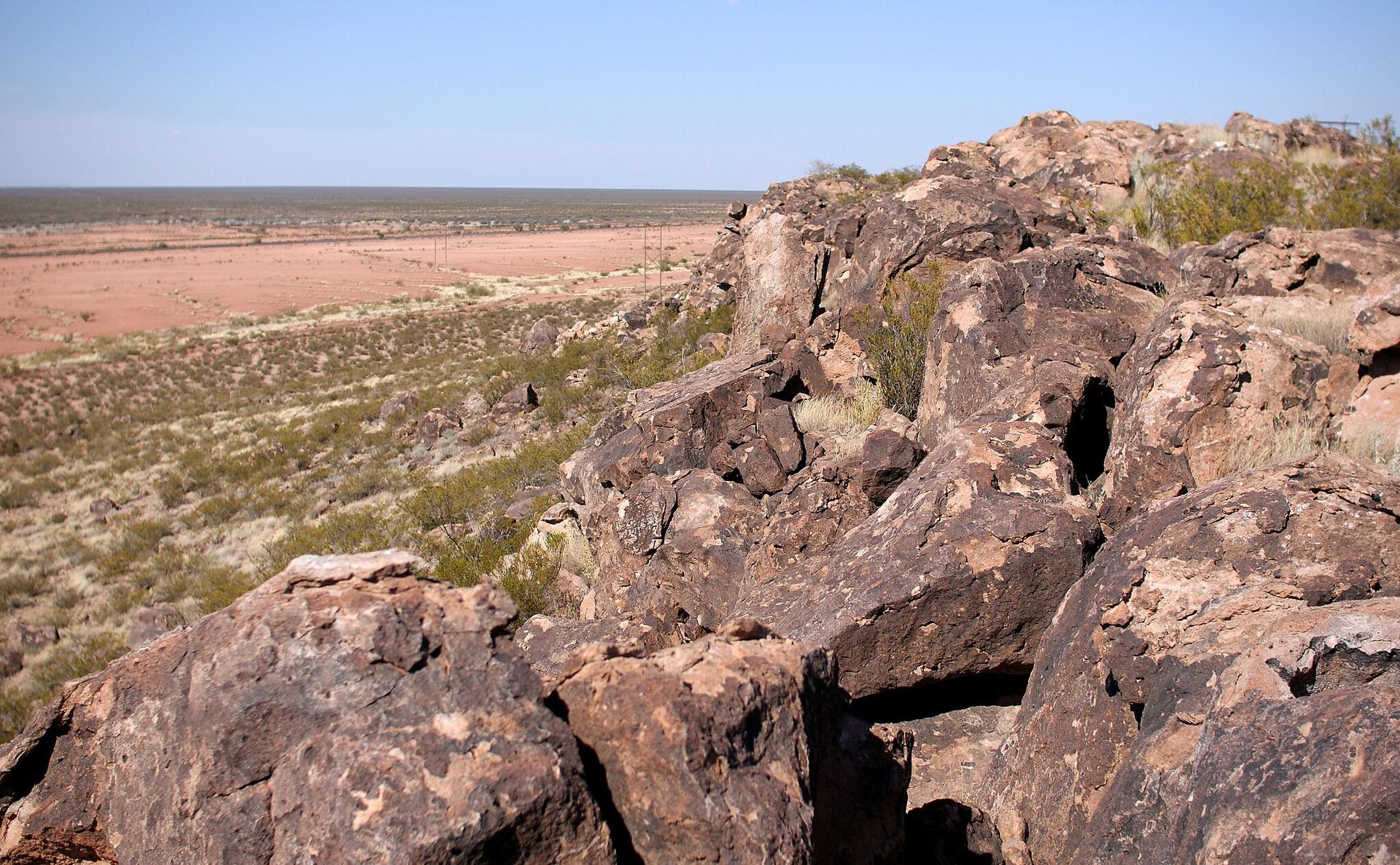 Sunlit rocky outcrop overlooks a wide, arid desert plain with sparse plants.