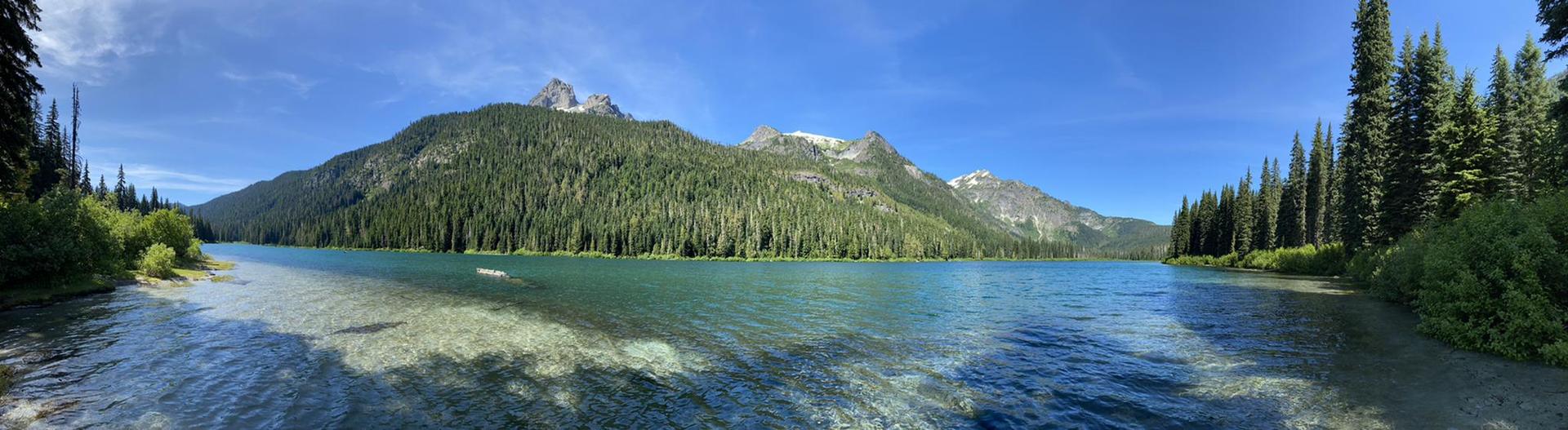 Panoramic view of a clear turquoise lake surrounded by forested mountains under a blue sky.