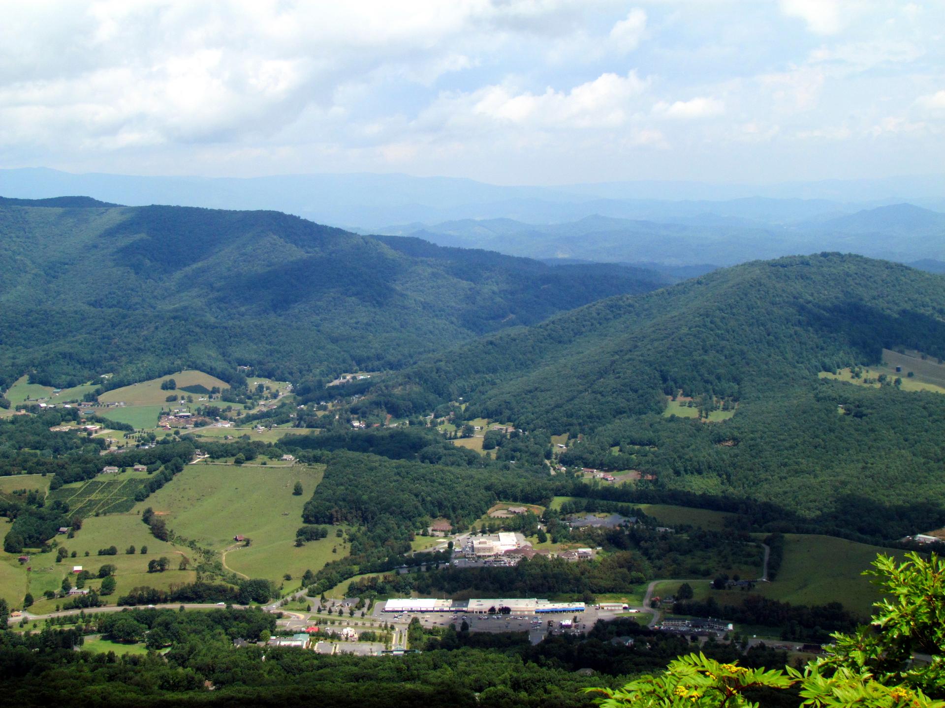 Green valley with buildings nestled among forested mountains under a cloudy sky.