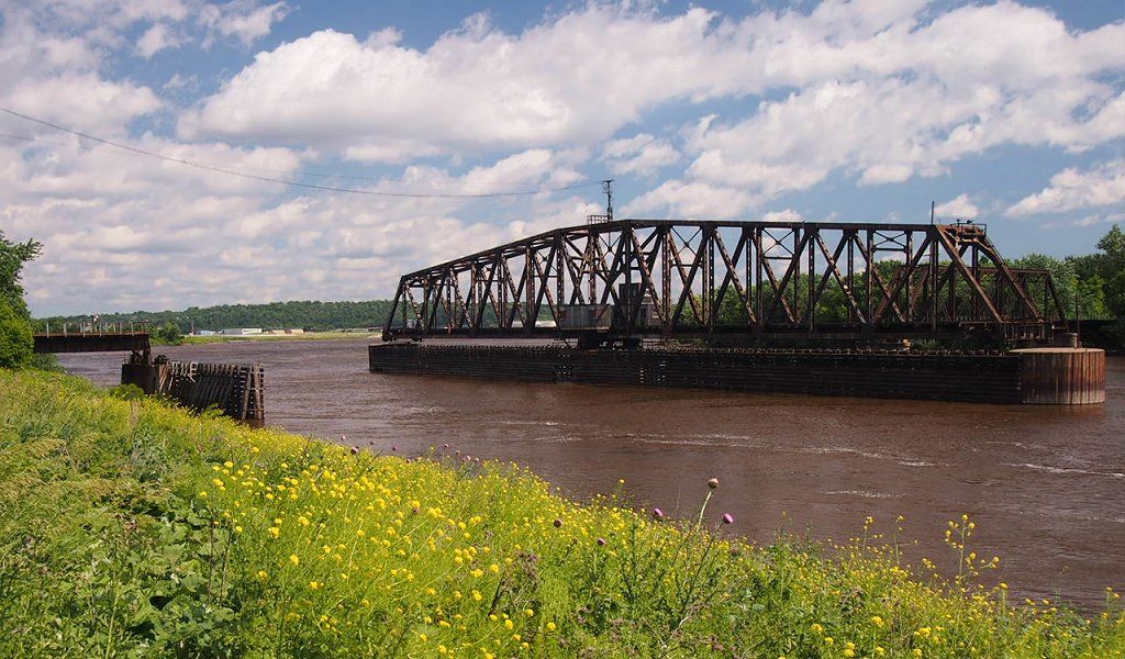 A large metal truss bridge spans a wide river under a cloudy sky. - River to River Greenway