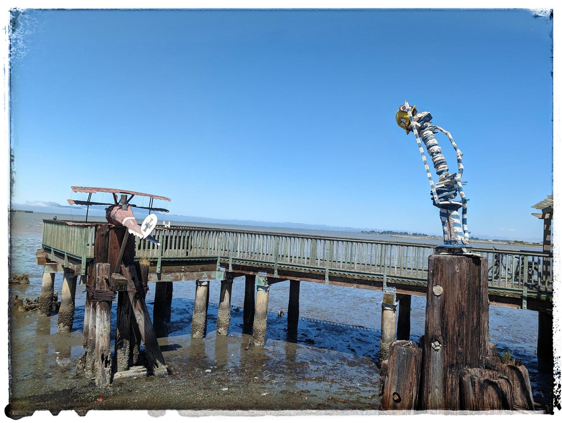 Wooden pier with two metal sculptures over calm water on a sunny day.