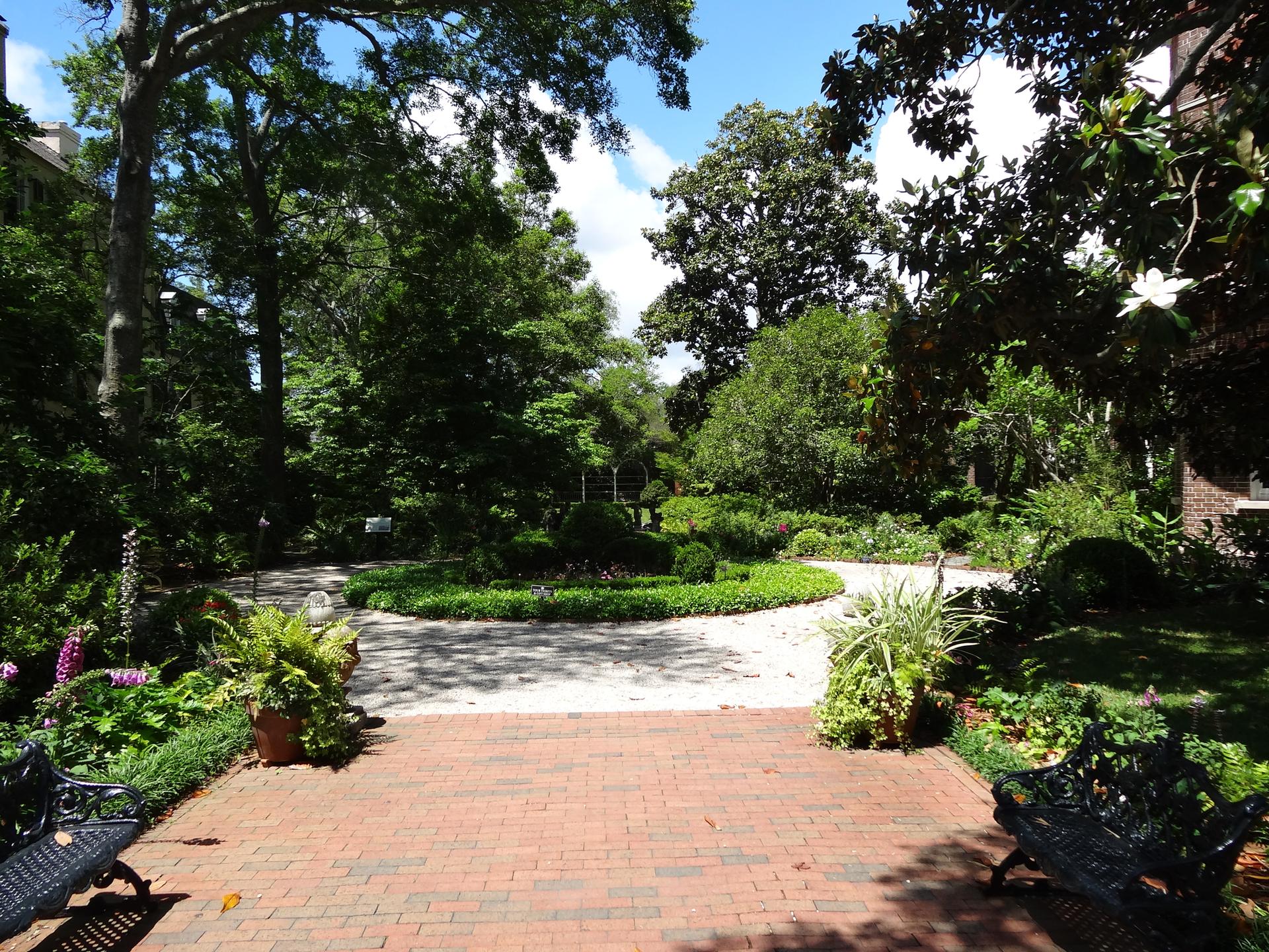 A sunny brick patio overlooks a lush, manicured garden with benches.
